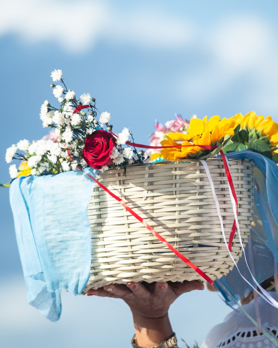 Mulher de blusa branca rendada levando nas mãos, acima da cabeça, um balaio de palha com rosas, girassóis, flores brancas, tule azul e fitas, contra o céu azul, a caminho do mar no Rio Vermelho, em Salvador, no dia de Iemanjá.