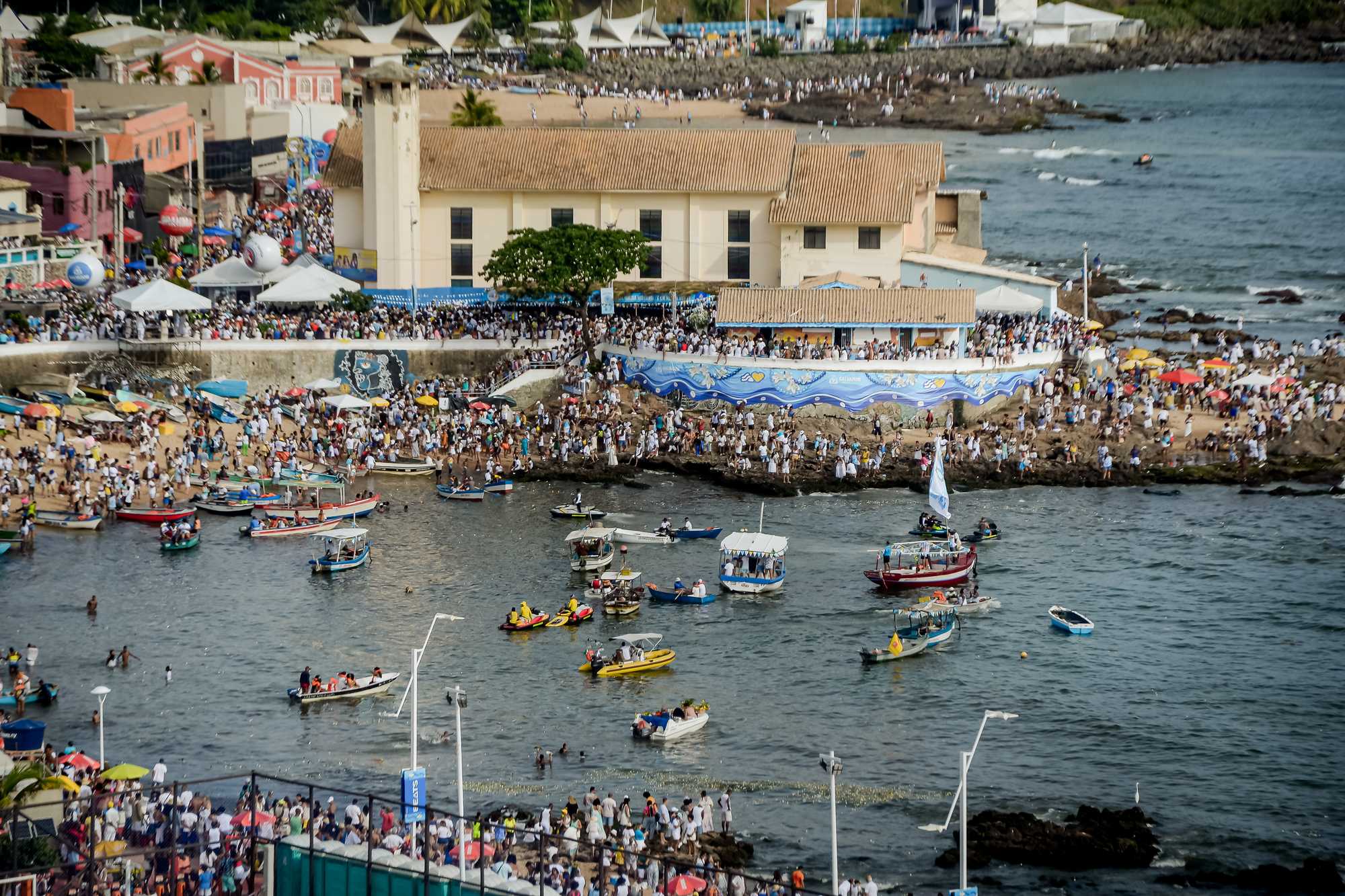 Vista do alto do Rio Vermelho, em Salvador, no 2 de fevereiro, com a praia, as pedras e o entorno da Casa de Iemanjá, de muro pintado de azul, tomados de gente vestida de branco, e dezenas de barcos na enseada.