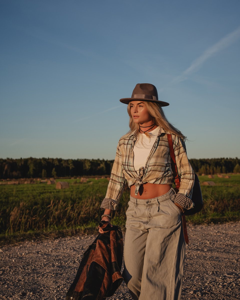 Mulher de chapéu de feltro, camisa xadrez amarrada na cintura e calça jeans larga caminhando numa estrada de terra ao pôr do sol, com uma jaqueta de couro na mão.