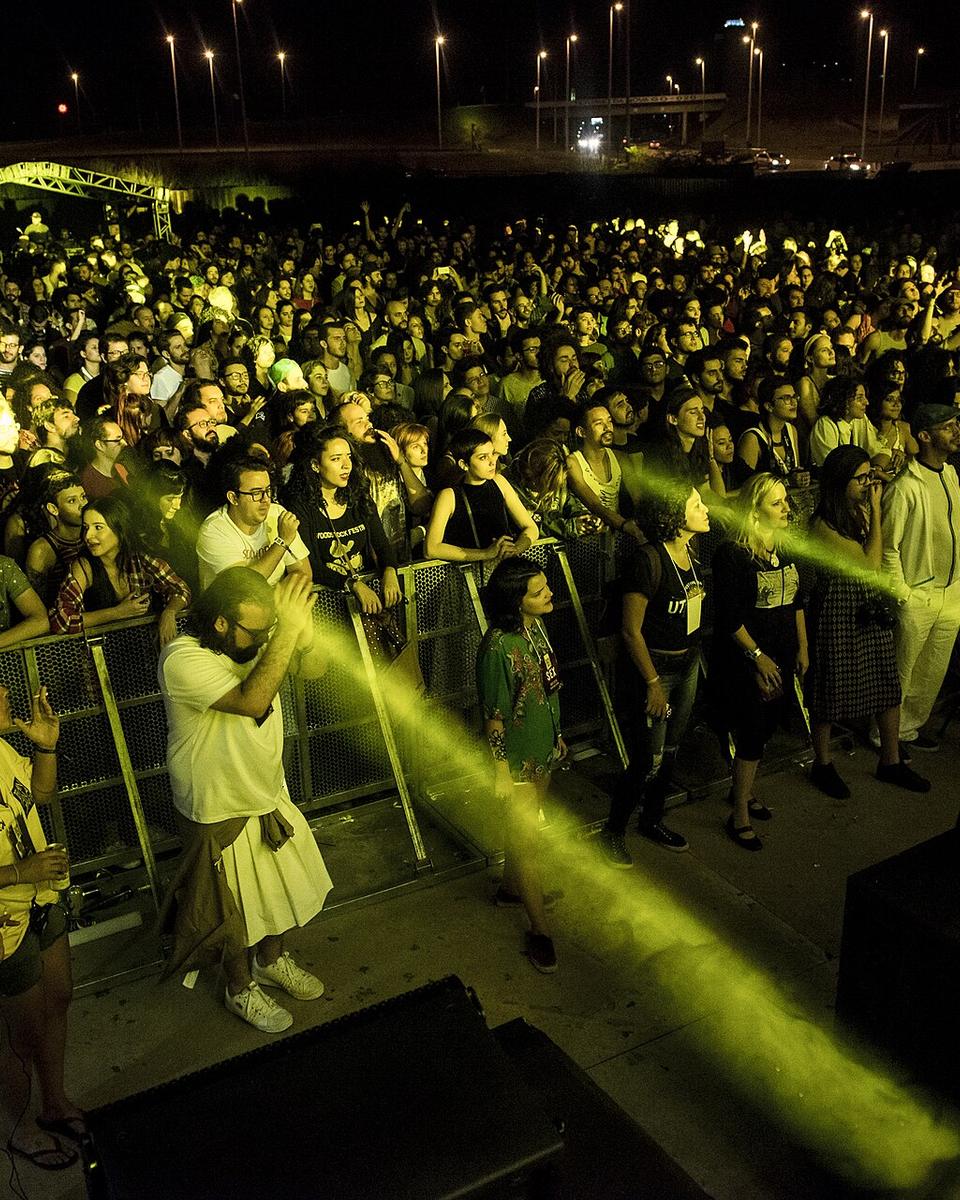 Público jovem encostado na grade em frente ao palco do Festival Bananada, em Goiânia, à noite, iluminado por um feixe de luz amarela, com gente de camiseta, vestido e mochila, alguns filmando com o celular.