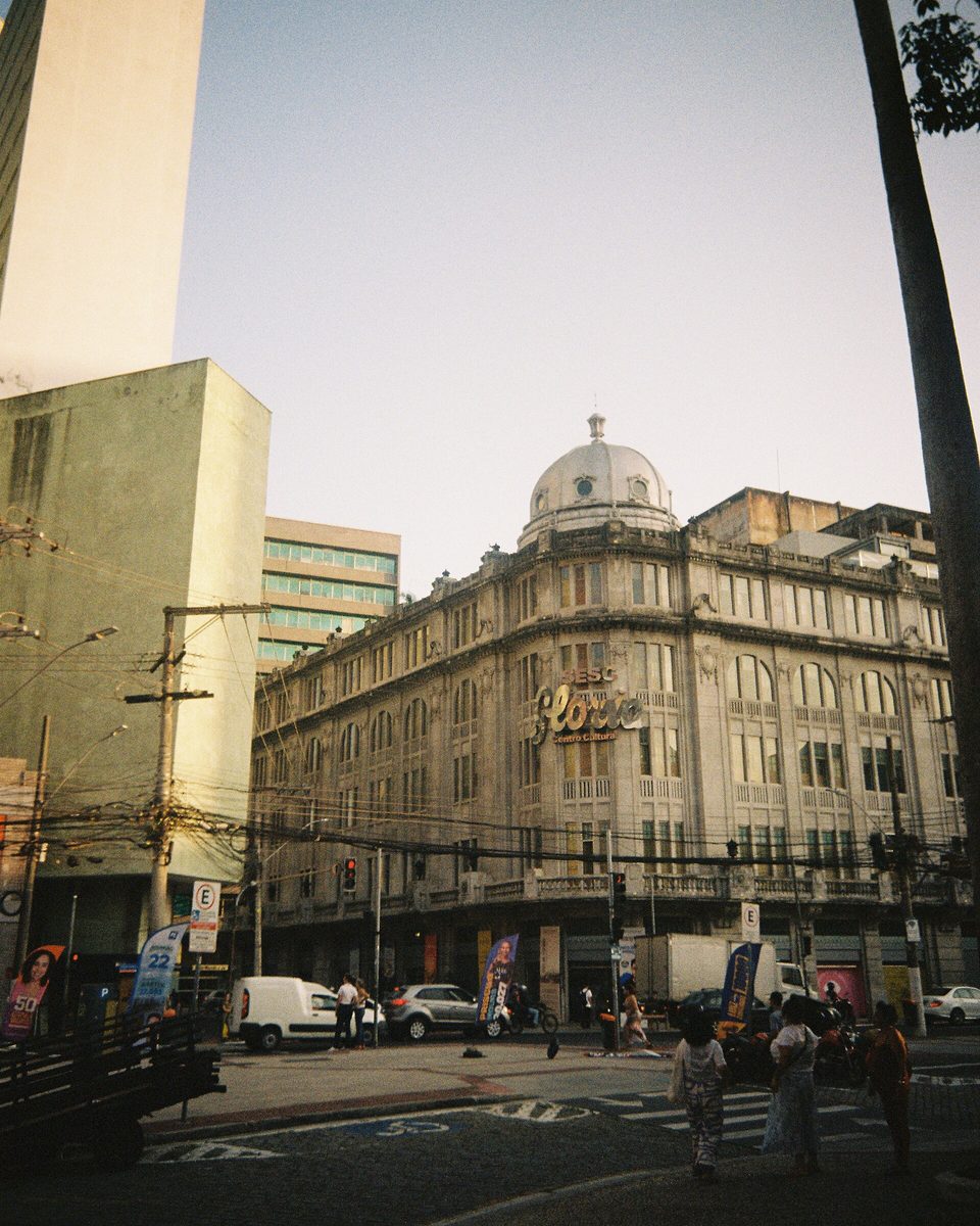 Esquina do Centro Cultural Sesc Glória, no Centro de Vitória, no fim da tarde, com pedestres atravessando a faixa e o letreiro do Glória aceso na fachada.