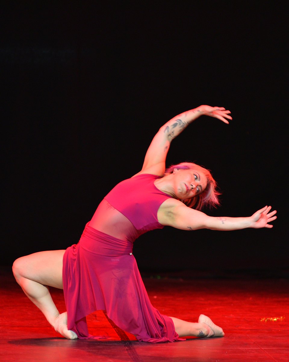 Bailarina adulta de vestido vermelho, com tatuagens nos braços, ajoelhada num palco iluminado de vermelho, inclinando o corpo para o lado com os braços abertos, numa apresentação do Festival de Dança de Joinville.