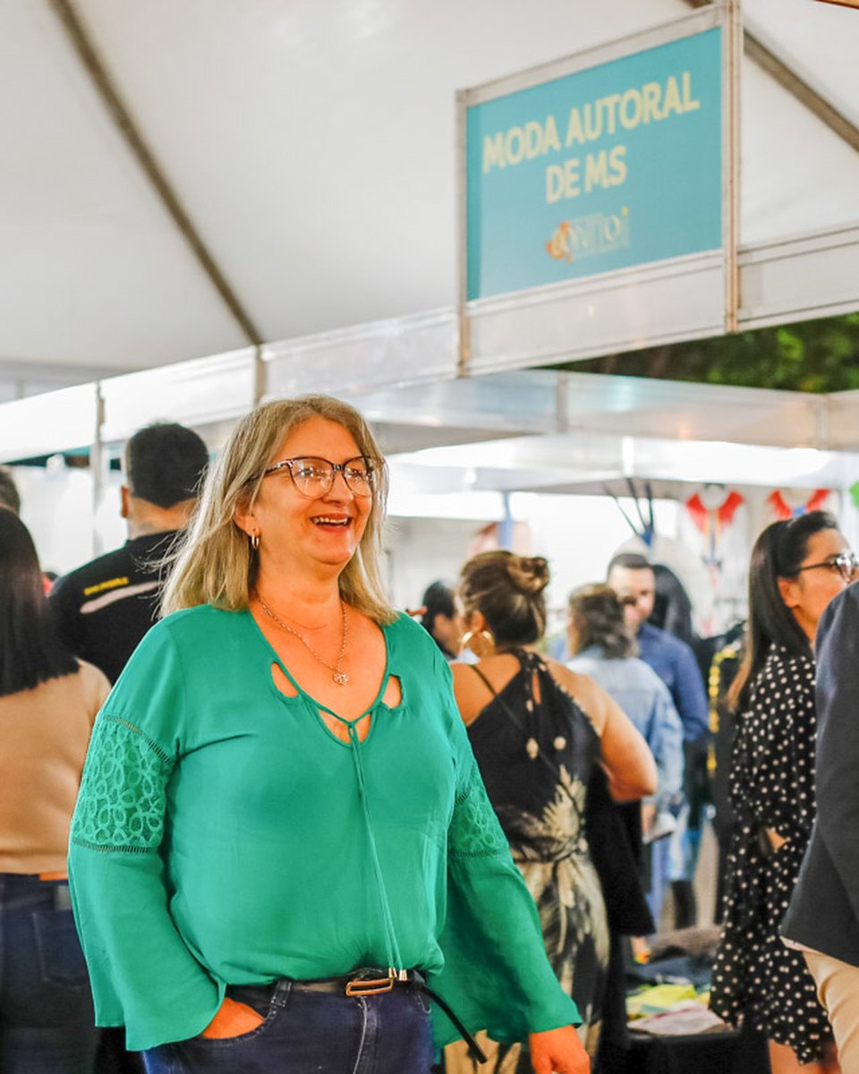 Visitantes circulando entre as barracas da tenda de moda autoral do Festival de Inverno de Bonito, no fim da tarde, com uma mulher de blusa verde sorrindo em primeiro plano.