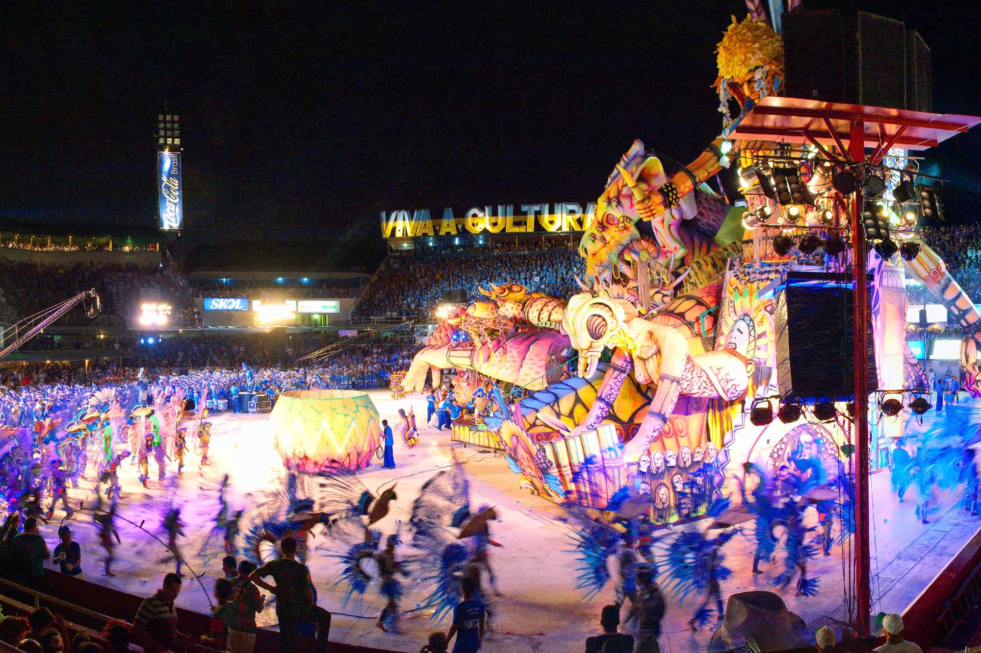 Vista geral do Bumbódromo de Parintins durante uma apresentação noturna, com alegorias gigantes iluminadas na arena, brincantes dançando na pista e as arquibancadas cheias dos dois lados.
