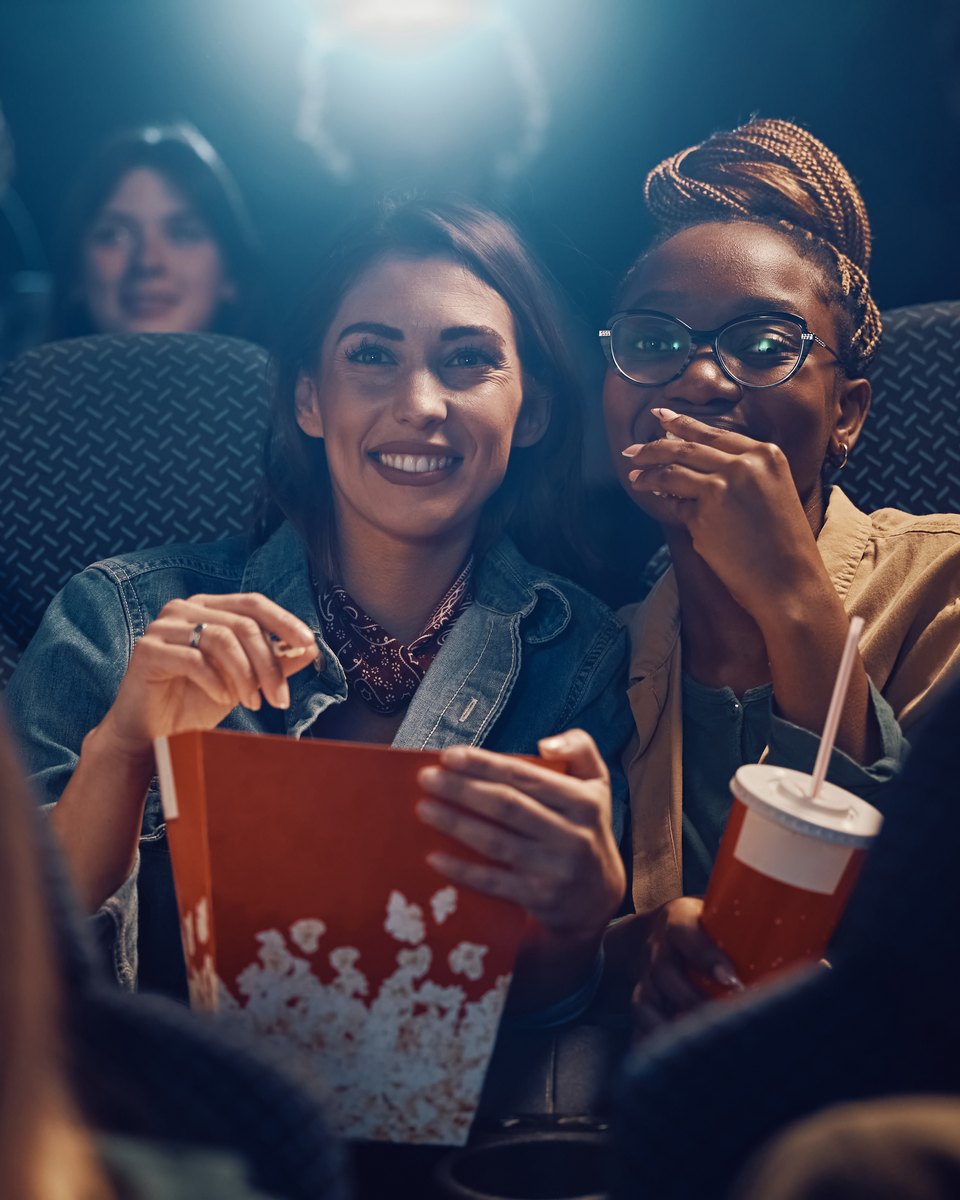 Duas amigas sorrindo numa sala de cinema escura, uma delas segurando um balde de pipoca, entre outros espectadores sentados nas poltronas.