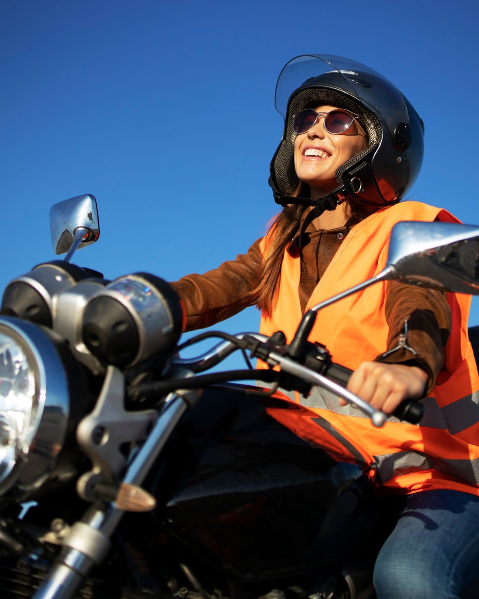 Mulher sorrindo de capacete aberto, óculos escuros e colete refletivo laranja, pilotando uma moto preta com baú traseiro, sob céu azul.