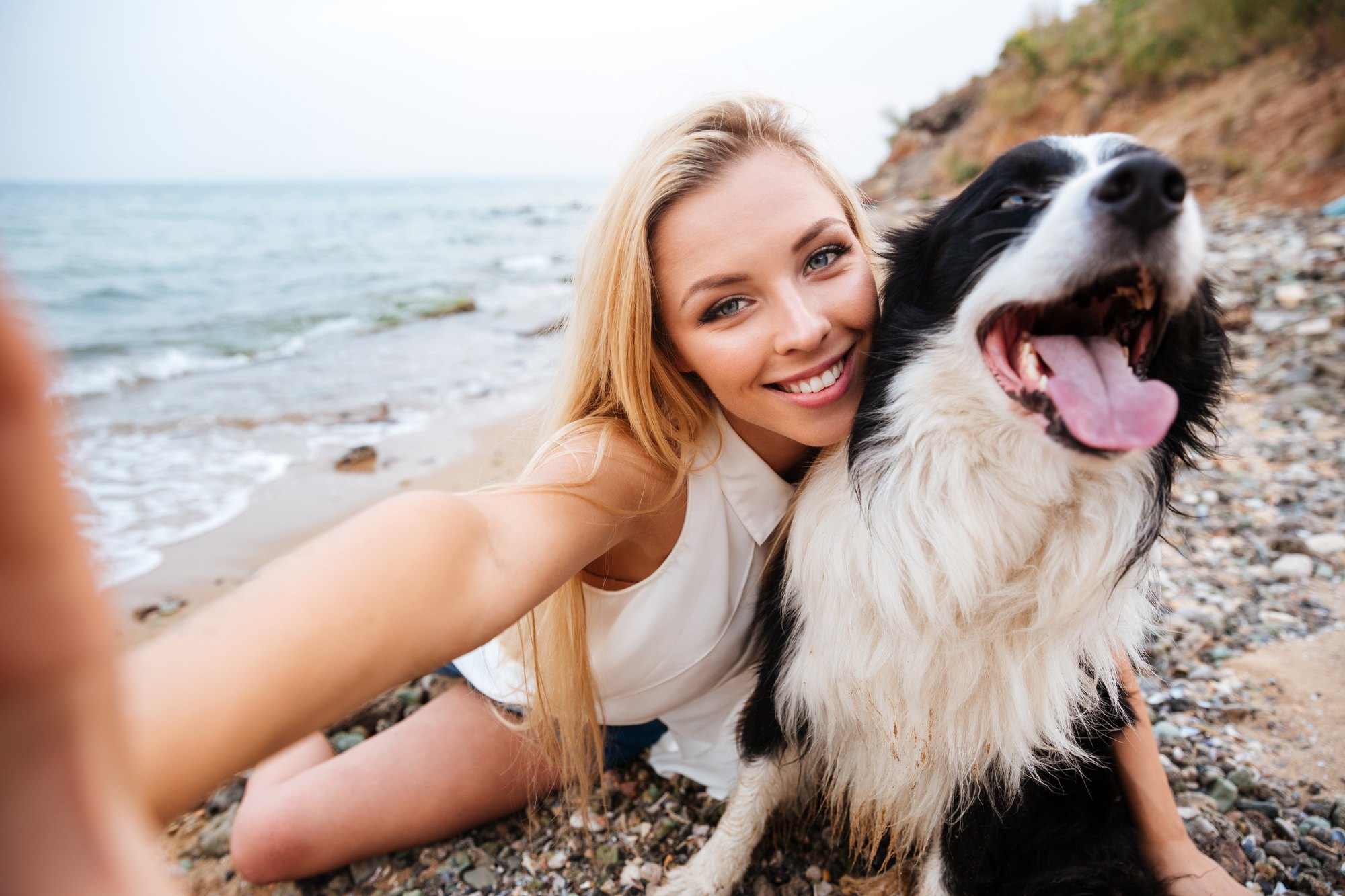 Mulher loira sorrindo e fazendo uma selfie abraçada a um border collie preto e branco de língua de fora, sentada numa praia de pedras à beira-mar.