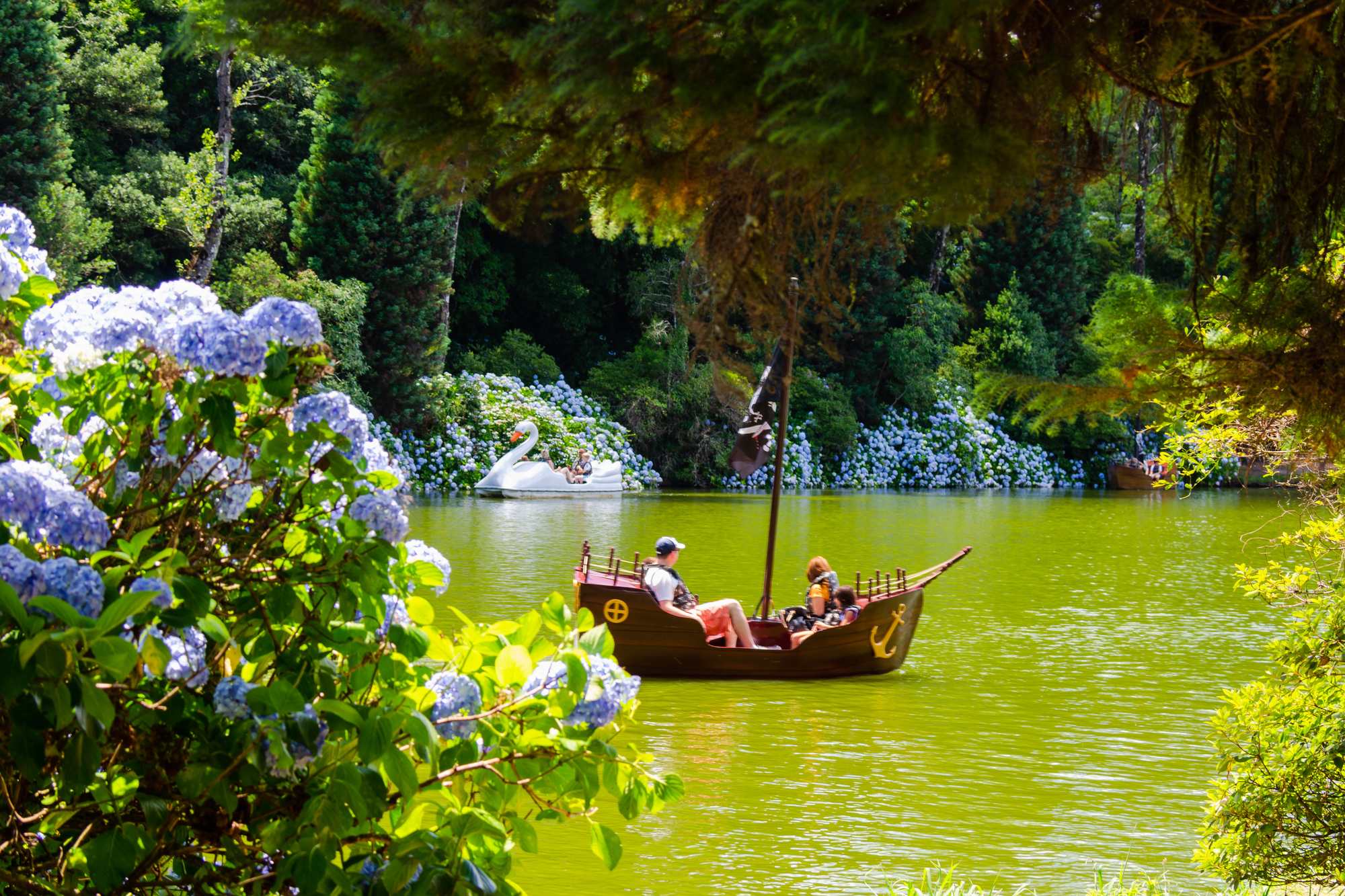 Turistas num pedalinho em forma de barco a vela no Lago Negro, em Gramado, com um pedalinho de cisne ao fundo e hortênsias azuis floridas na margem.