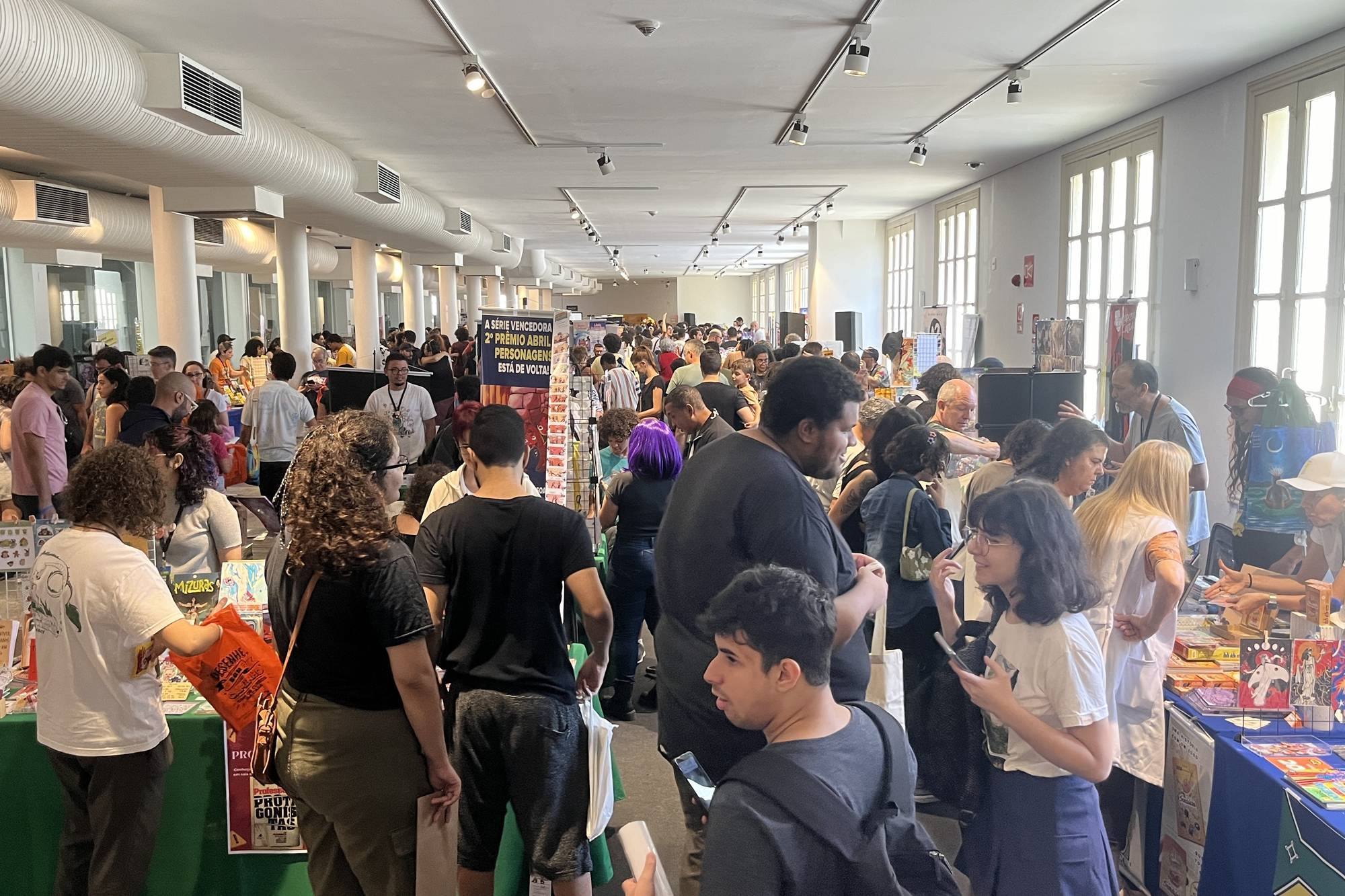 Corredor lotado da feira de quadrinistas do FIQ BH 2024, no Minascentro, com visitantes folheando gibis e livros nas mesas de artistas, muitos de sacola na mão, sob janelões e luzes do salão.