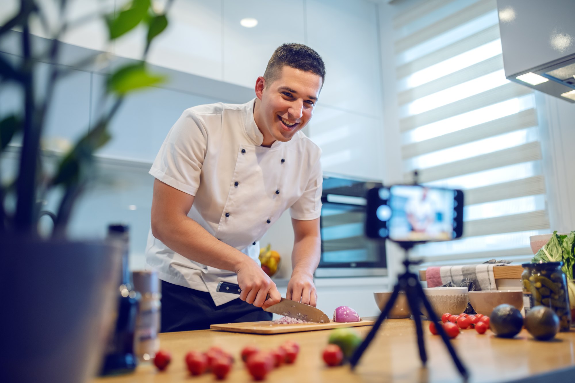 Chef de dólmã branco sorrindo enquanto pica cebola numa bancada de cozinha, gravando a si mesmo com um celular num tripé à frente.