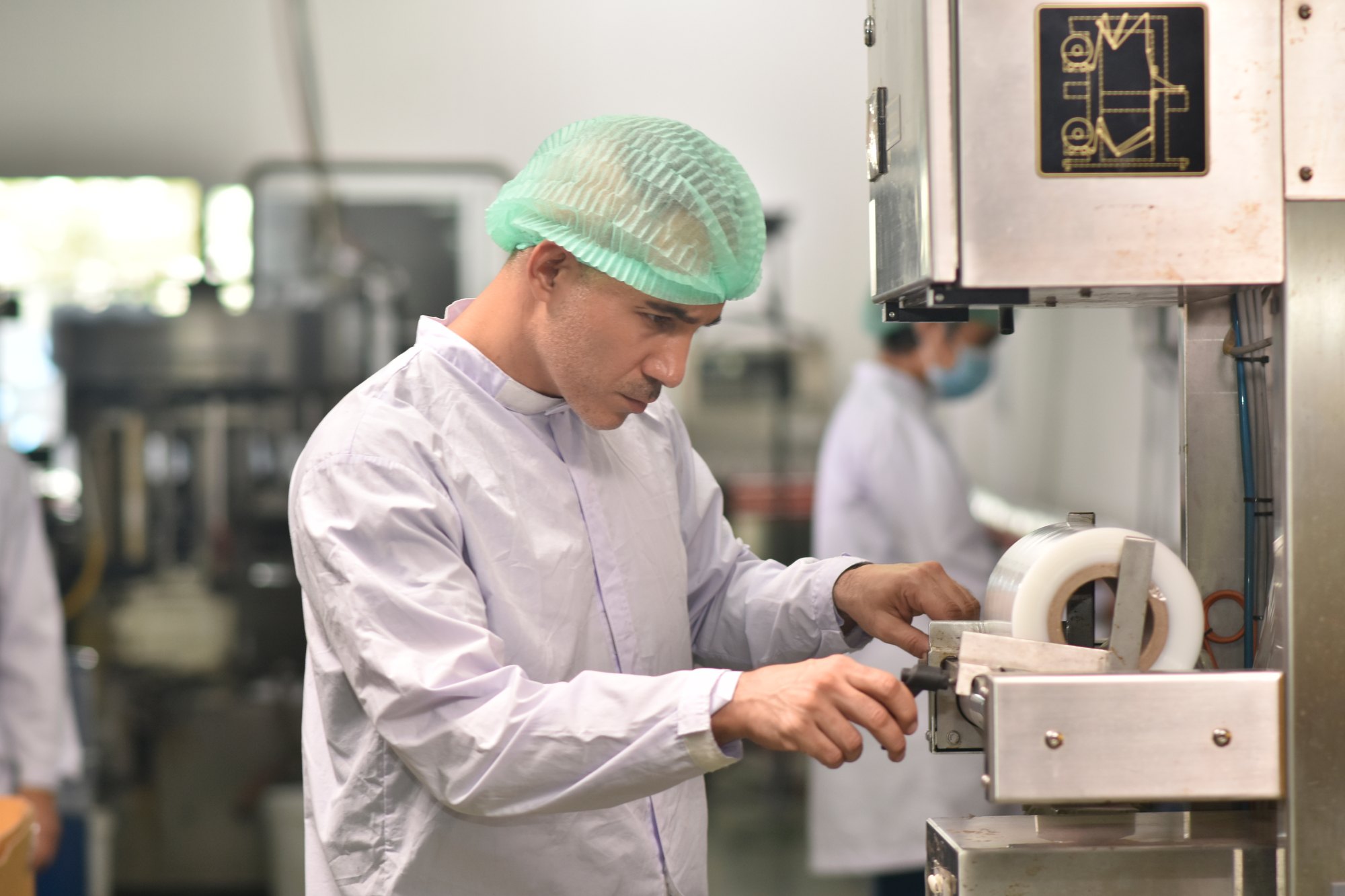 Operador de touca descartável e jaleco branco ajustando uma bobina de filme numa máquina de embalagem, dentro de uma fábrica de alimentos.