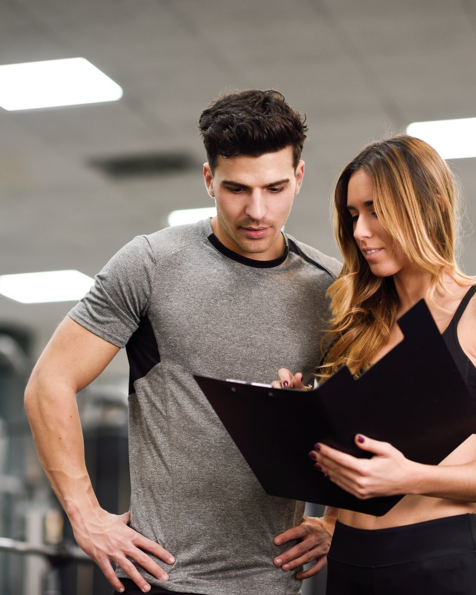 Personal trainer de camiseta cinza e aluna de top preto olhando juntos uma prancheta, em pé numa academia com aparelhos de musculação fora de foco ao fundo.