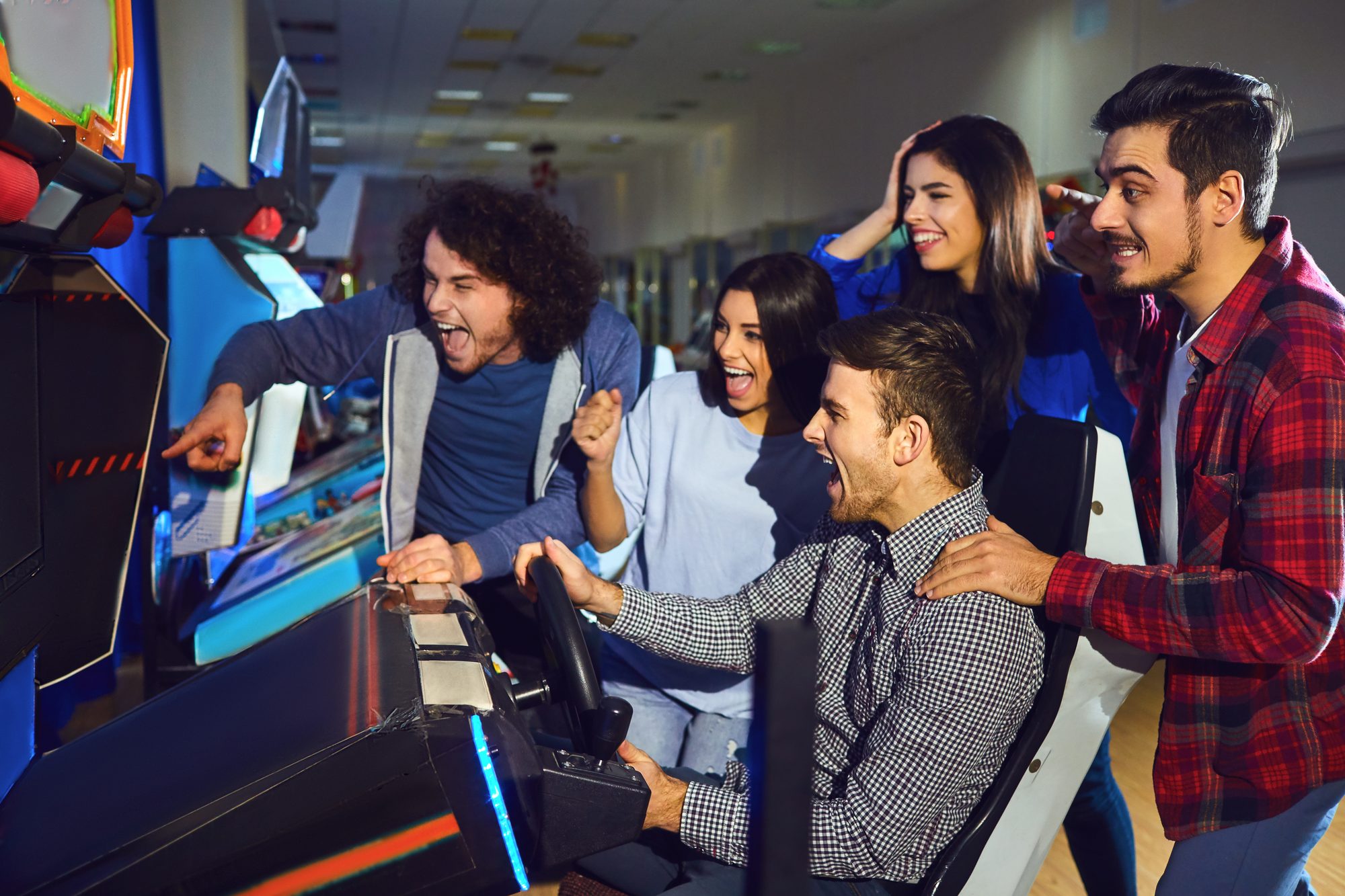 Grupo de cinco jovens rindo e torcendo em volta de uma máquina de fliperama de corrida, num salão de jogos com luz azul.
