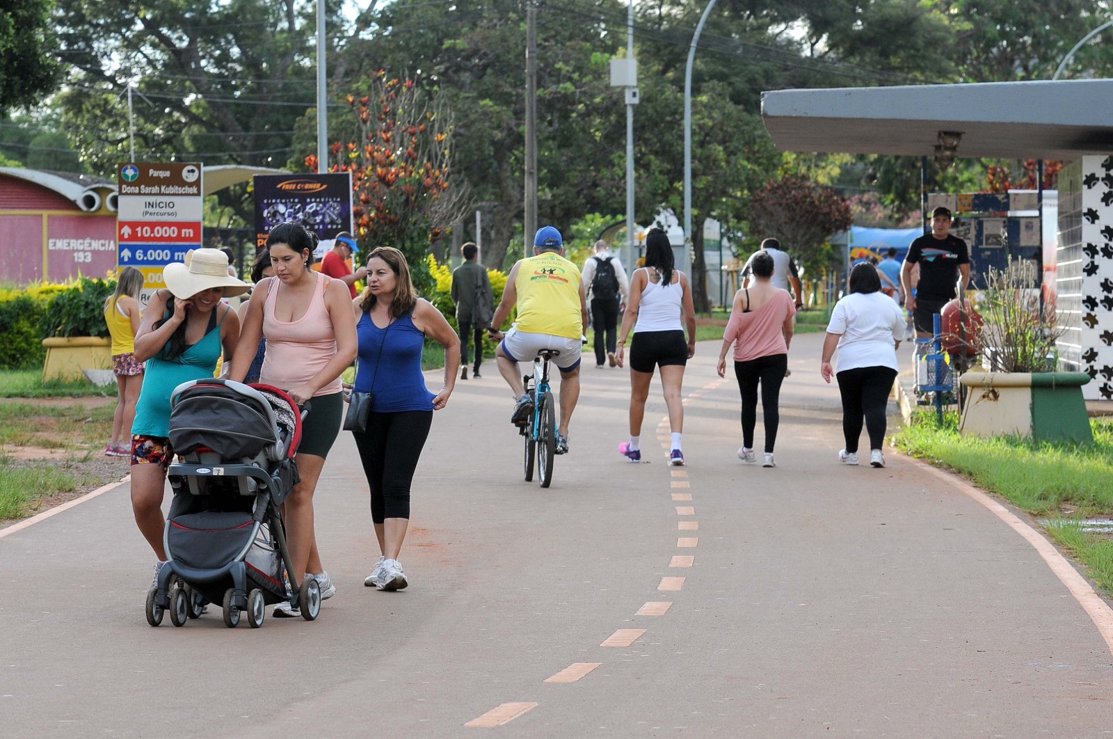 Pessoas caminhando e pedalando na pista do Parque da Cidade Dona Sarah Kubitschek, em Brasília, com duas mulheres empurrando um carrinho de bebê em primeiro plano, placa de percurso de 10 mil e 6 mil metros à esquerda e árvores ao fundo.