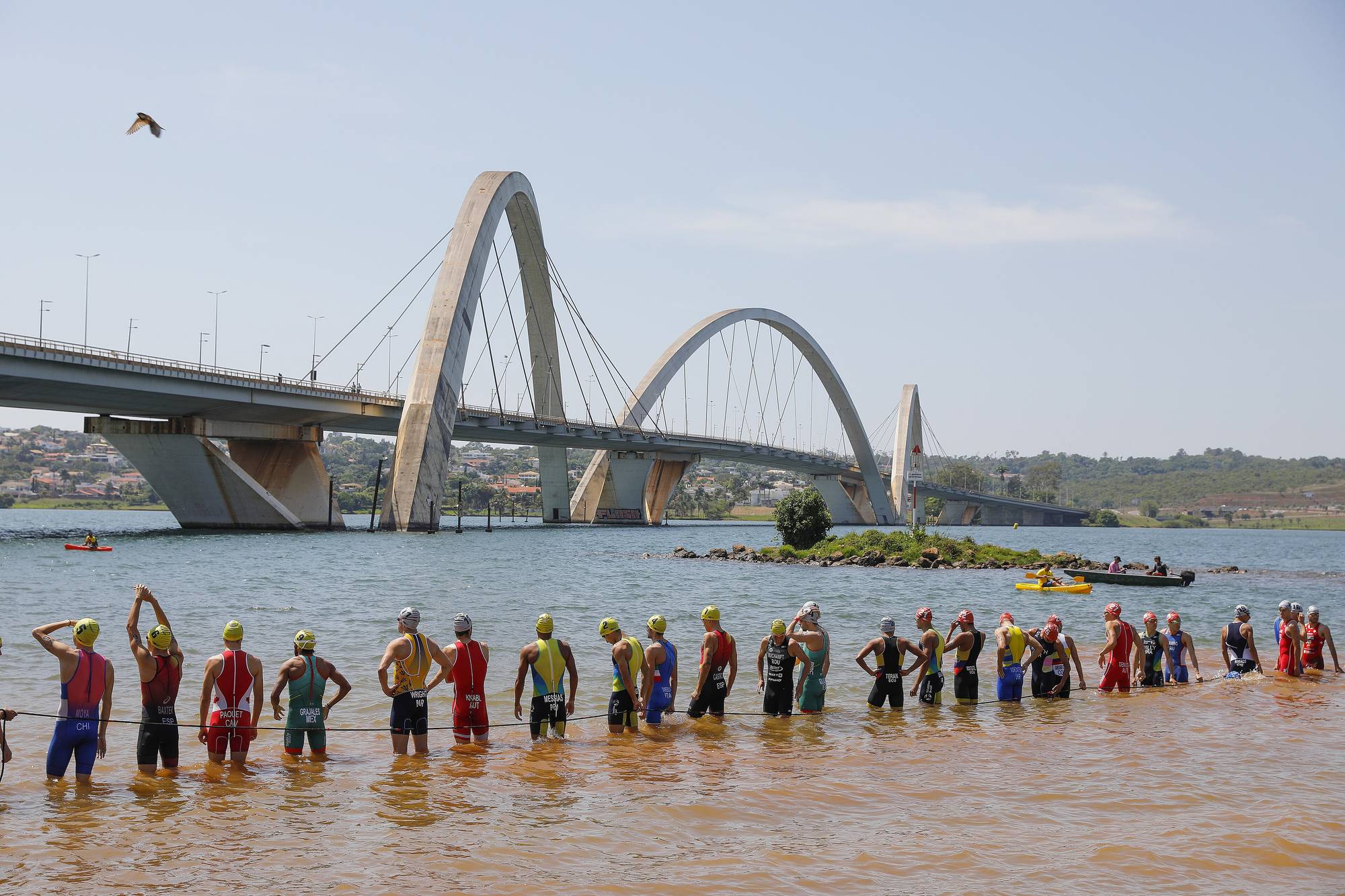 Fila de triatletas de touca colorida com a água do Lago Paranoá pela cintura, antes da largada da natação, com a ponte de três arcos de Brasília ao fundo, na etapa de Brasília da Copa do Mundo de Triathlon de 2023.