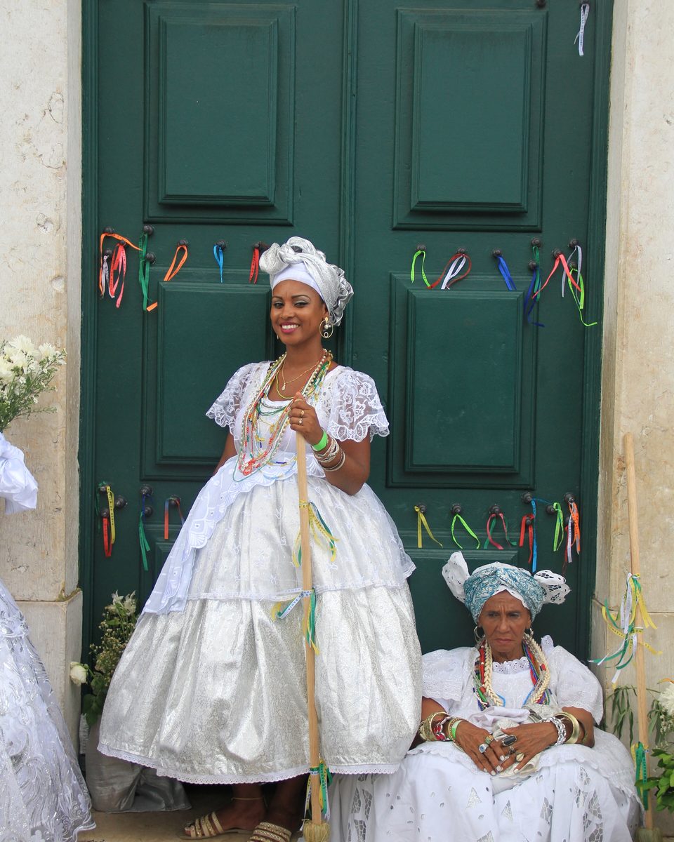 Duas baianas vestidas de branco, com torsos, colares de contas e vassouras enfeitadas de fitas, diante da porta verde de uma igreja com fitinhas coloridas amarradas, durante a Lavagem do Bonfim, em Salvador.
