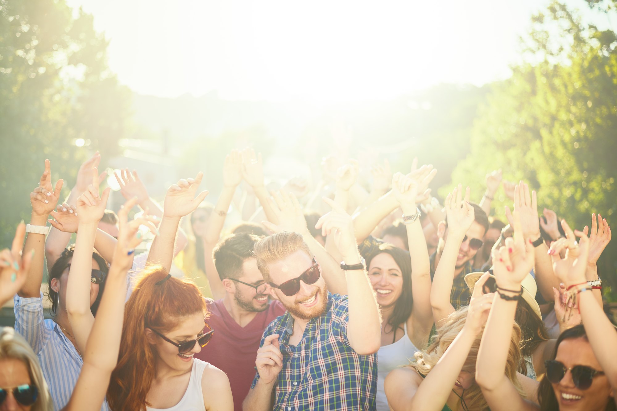 Público jovem de óculos escuros dançando com os braços para cima num festival de música ao ar livre, num dia de sol forte.
