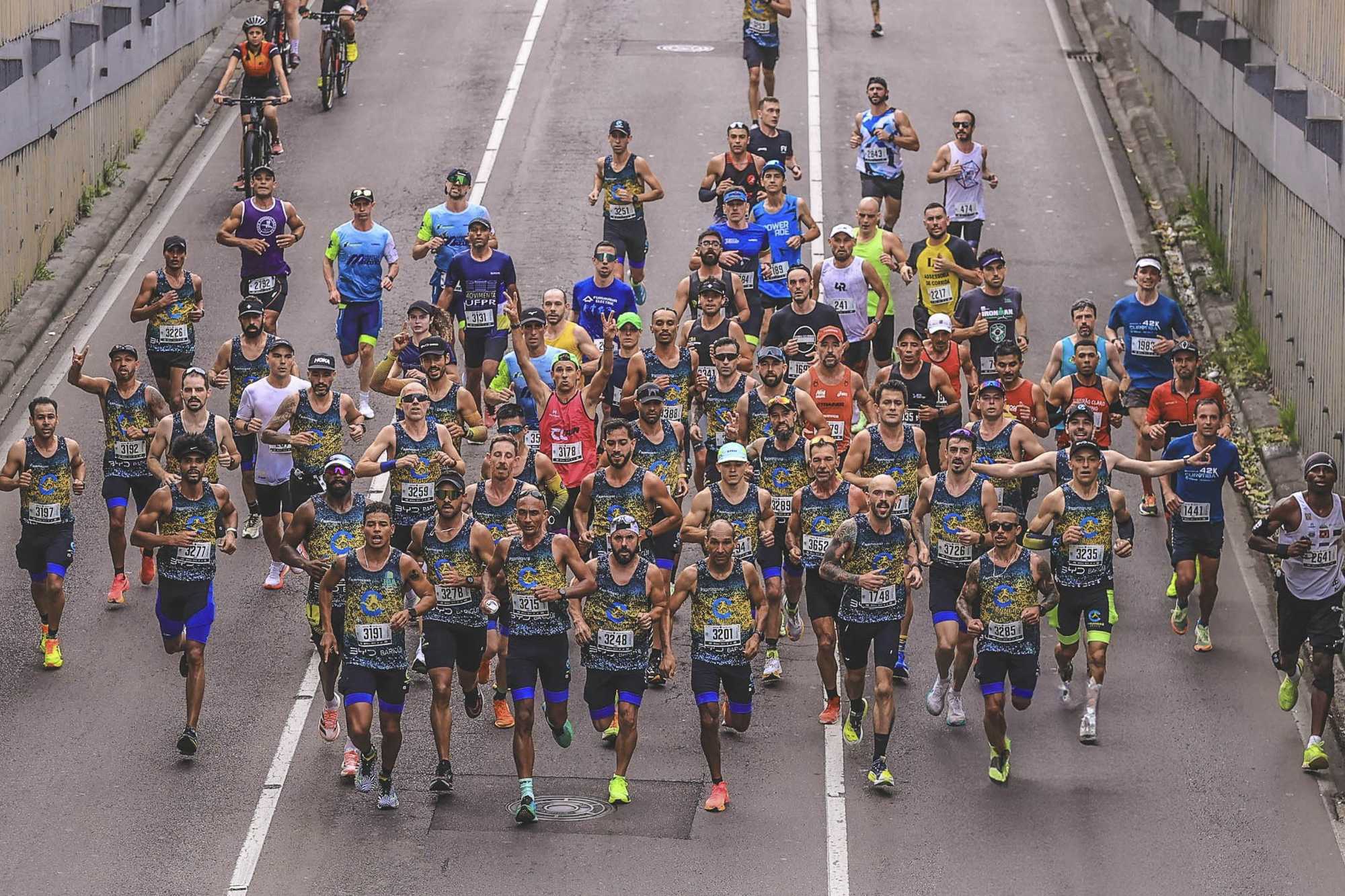 Pelotão de maratonistas com número de peito descendo uma trincheira de concreto durante a Maratona de Curitiba.