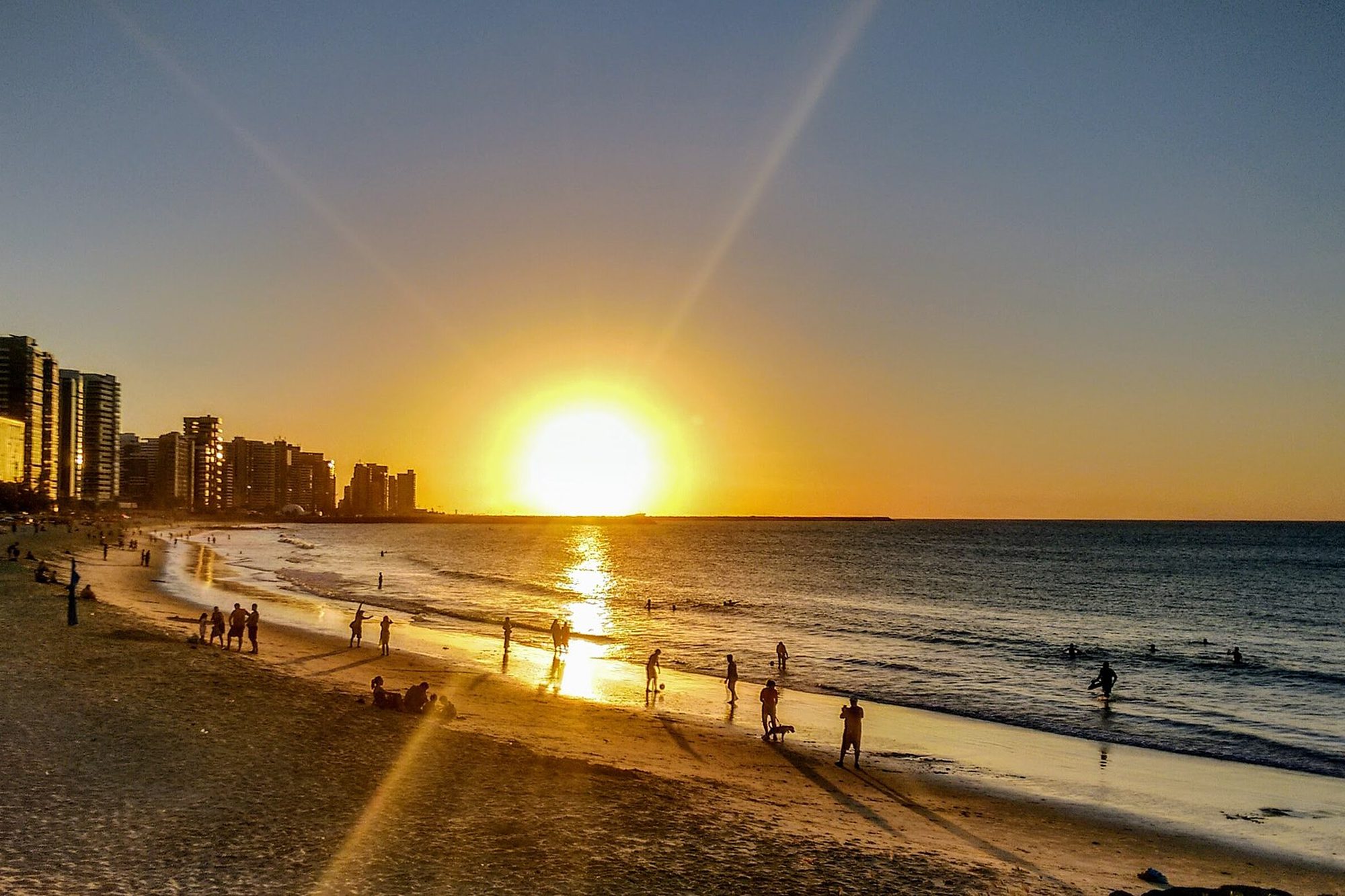 Sol baixo sobre o mar na Praia de Iracema, em Fortaleza, com pessoas caminhando e correndo na beira da água e os prédios da Beira-Mar ao fundo.