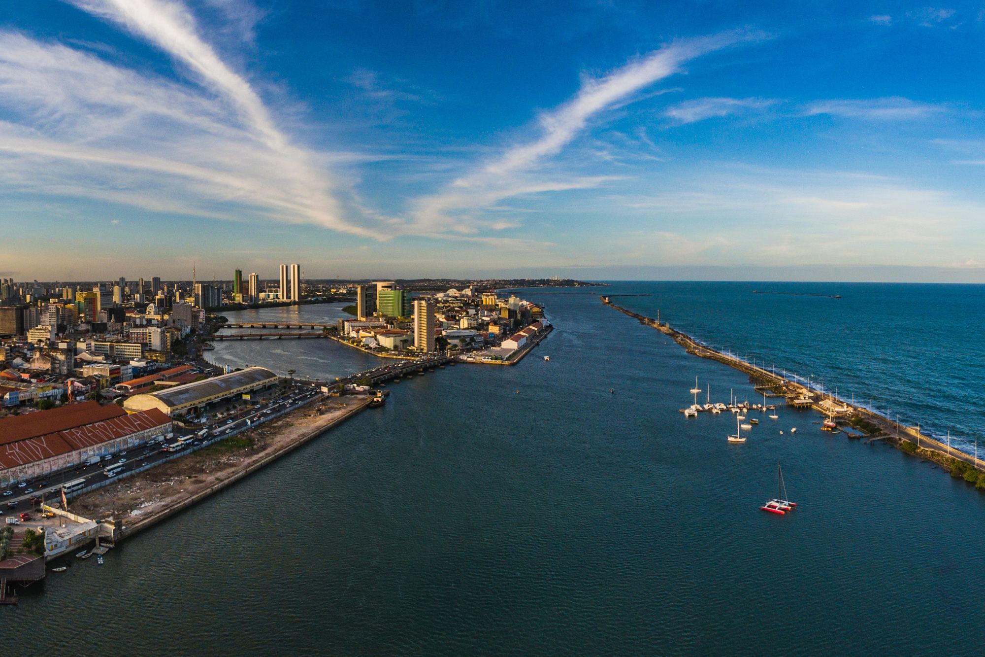 Vista aérea do porto do Recife ao entardecer, com os armazéns do cais e uma avenida movimentada à esquerda, pontes sobre o rio e os prédios do centro ao fundo, o molhe dos arrecifes com veleiros ancorados à direita e o mar aberto sob céu azul com nuvens finas.