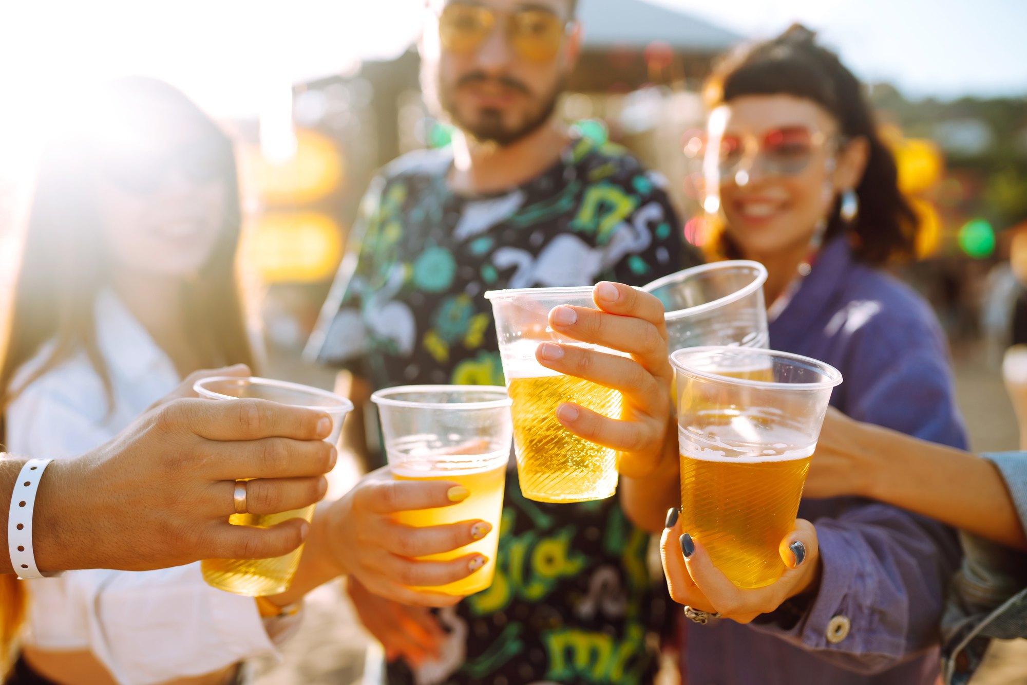 Grupo de amigos adultos brindando com copos de cerveja num festival ao ar livre, com luz de fim de tarde e barracas desfocadas ao fundo.