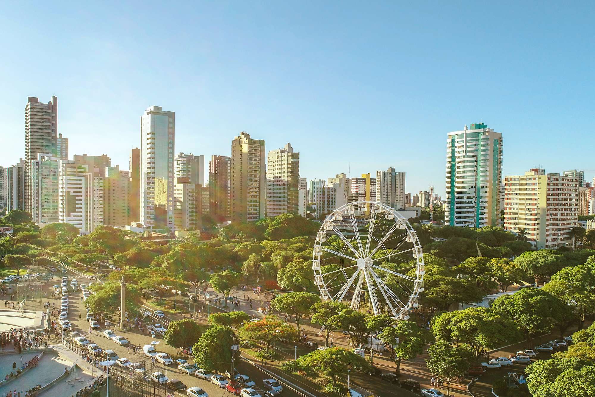 Vista do alto do centro de Maringá num fim de tarde de sol, com uma roda-gigante branca entre as copas das árvores, avenidas com carros, pessoas caminhando numa praça com chafariz e prédios ao fundo.