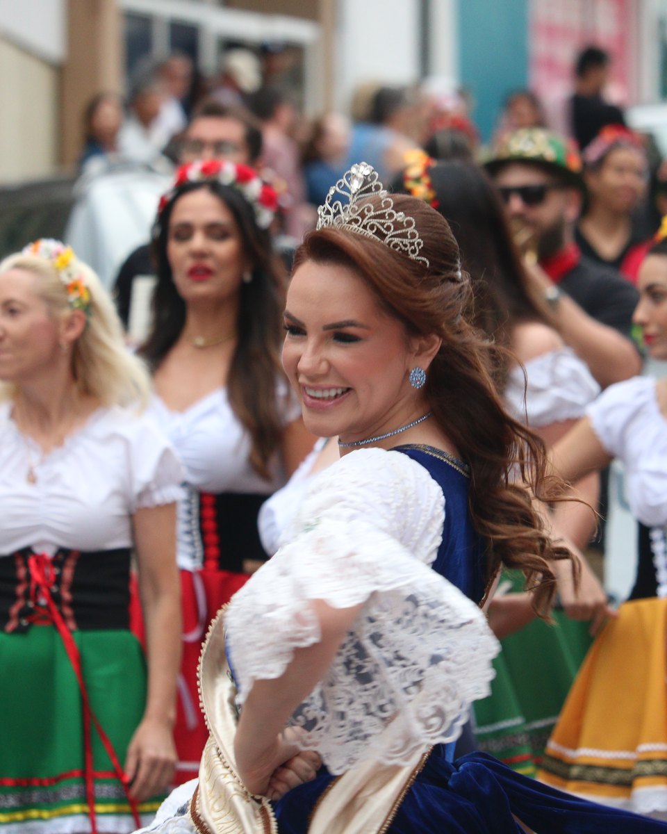 Mulher de tiara, faixa de majestade e vestido típico azul com mangas de renda branca dança sorrindo no desfile da Münchenfest, em Ponta Grossa, com participantes de trajes alemães coloridos e chapéus tiroleses atrás.