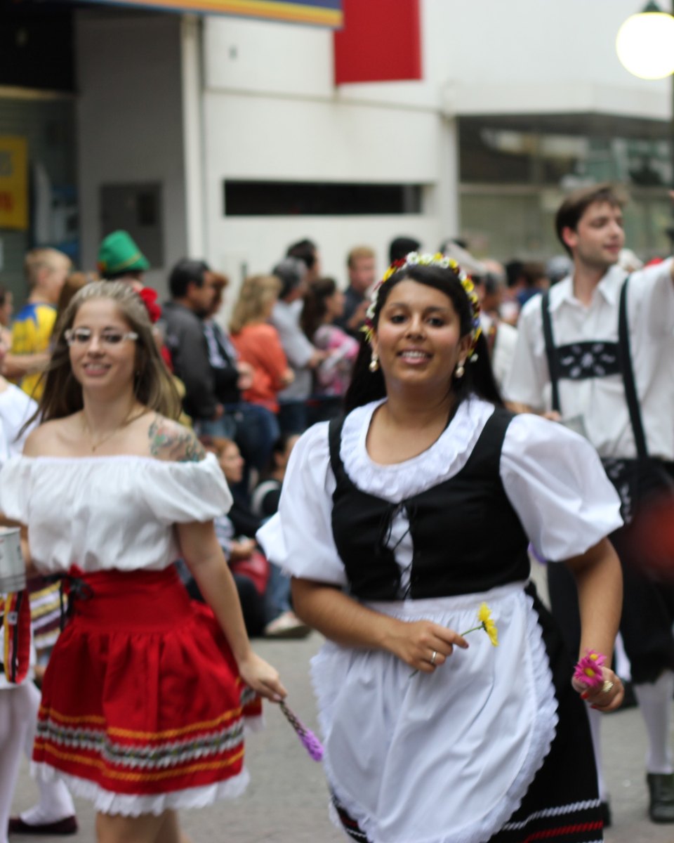 Jovem sorrindo com coroa de flores, blusa branca bufante, corpete preto e avental branco caminhando no desfile da Oktoberfest em Blumenau, ao lado de uma moça de saia vermelha e de rapazes de suspensório, com o público na calçada ao fundo.