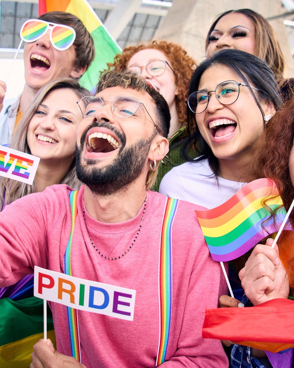 Grupo de amigos sorrindo numa selfie, com bandeiras de arco-íris, óculos coloridos e plaquinhas escritas Pride e Love, numa celebração do orgulho LGBT+.