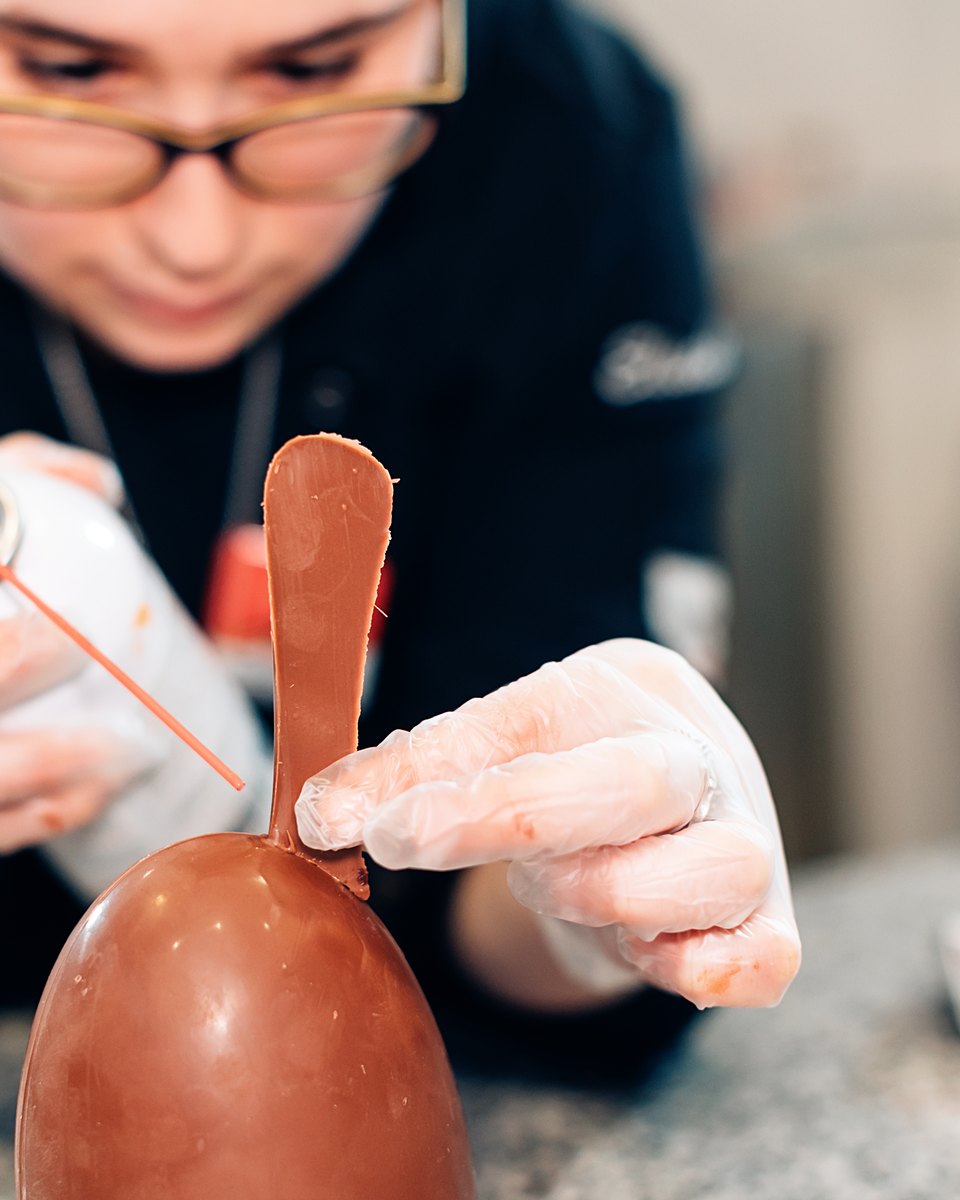 Confeiteira de luvas finalizando um ovo de chocolate com orelha de coelho numa bancada de ateliê.