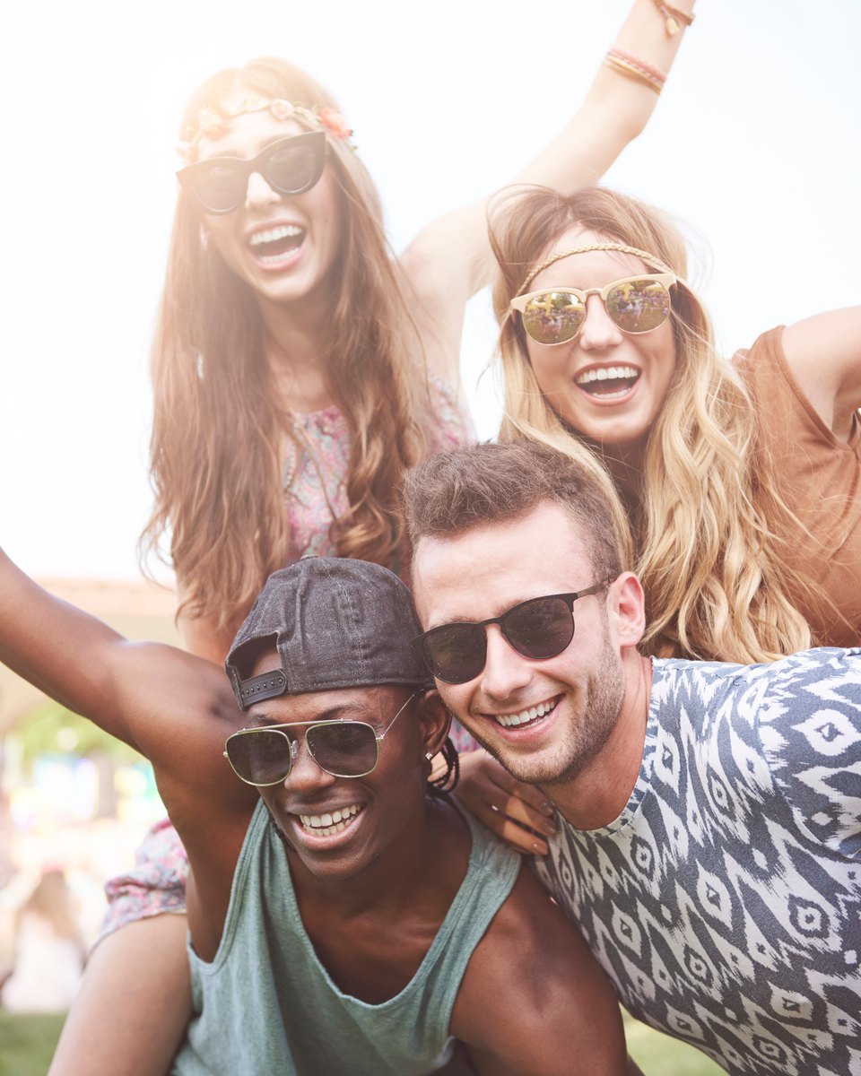 Quatro amigos sorrindo de óculos escuros num festival de música ao ar livre, com duas moças sentadas nos ombros de dois rapazes, num dia de sol.