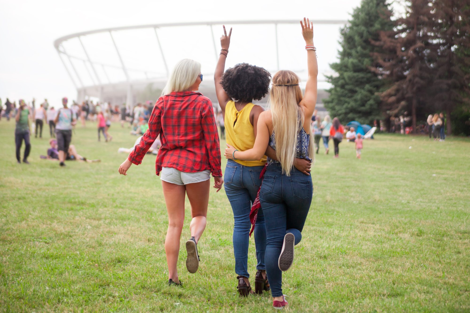 Três amigas de costas, abraçadas e com o braço para cima, caminhando num gramado de festival de música, com a estrutura de um estádio ao fundo.