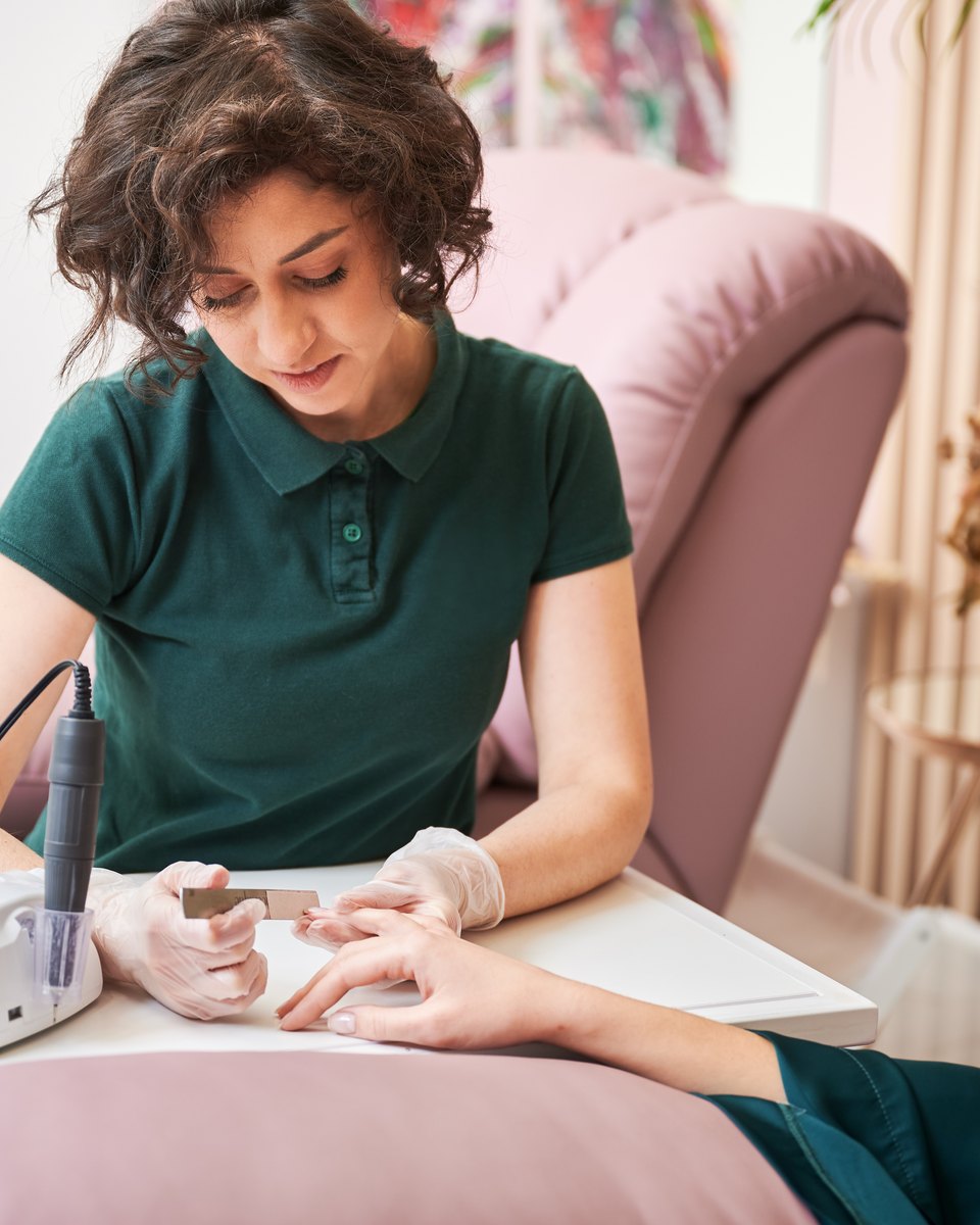 Manicure de camisa verde usando uma lixa elétrica nas unhas de uma cliente, sentada à mesa de um estúdio de unhas com poltrona rosa.