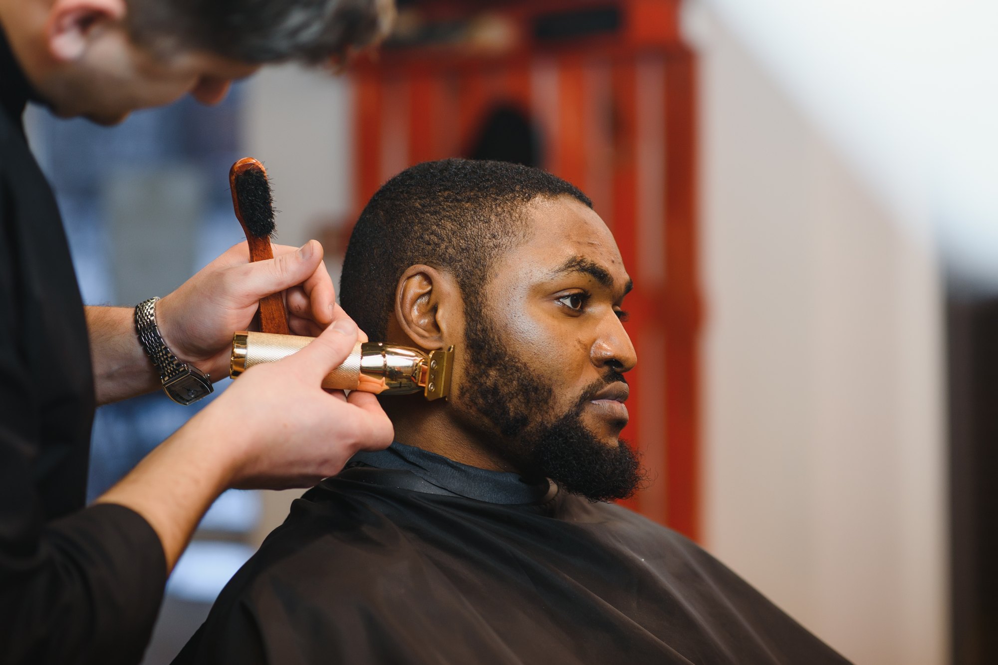 Barbeiro aparando com uma máquina dourada a barba de um cliente negro de capa preta, sentado na cadeira de uma barbearia.