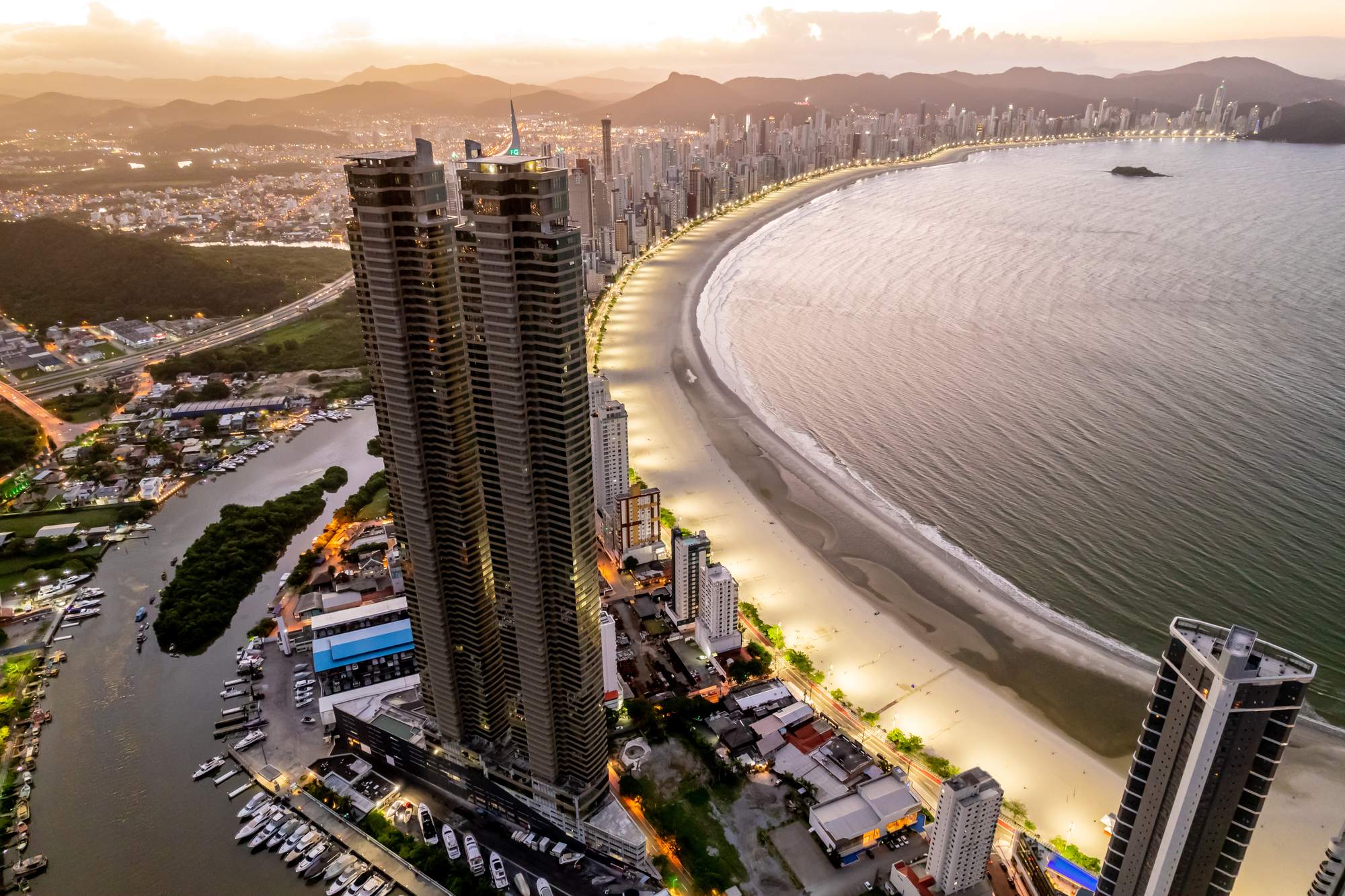 Vista aérea ao entardecer de Balneário Camboriú, com duas torres residenciais muito altas ao lado da marina do rio Camboriú e a Praia Central em curva, iluminada, cercada de prédios até o morro ao fundo.