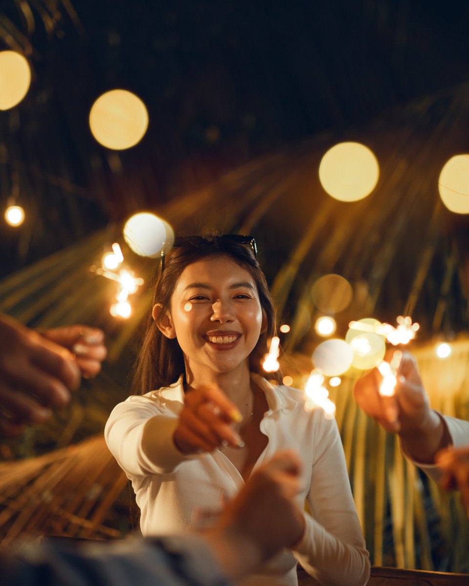 Grupo de amigos rindo e segurando estrelinhas acesas numa festa ao ar livre à noite, sob um varal de luzes.