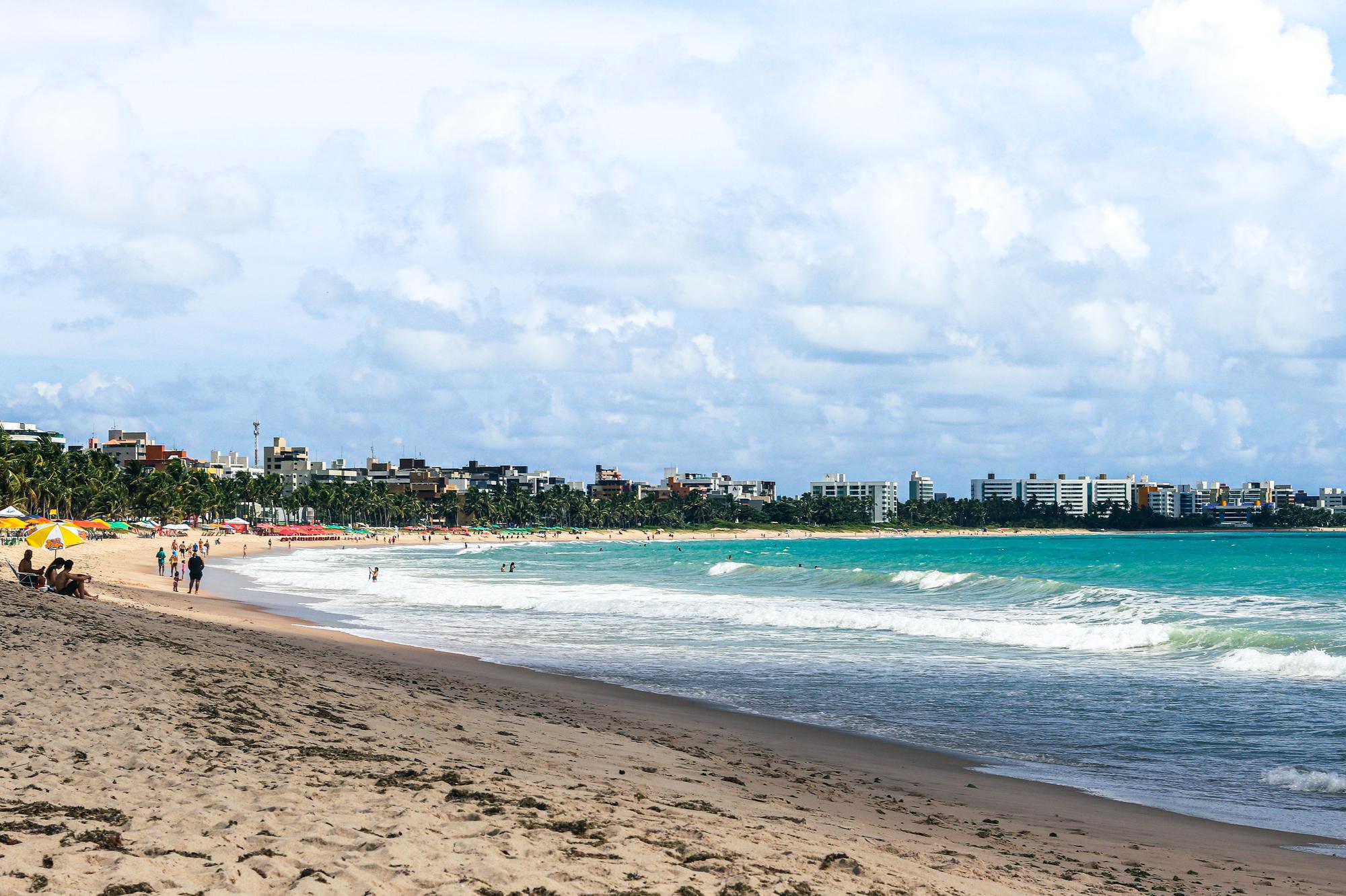 Praia de João Pessoa vista da areia, com guarda-sóis coloridos, banhistas na beira d'água e a faixa de prédios da orla ao fundo.