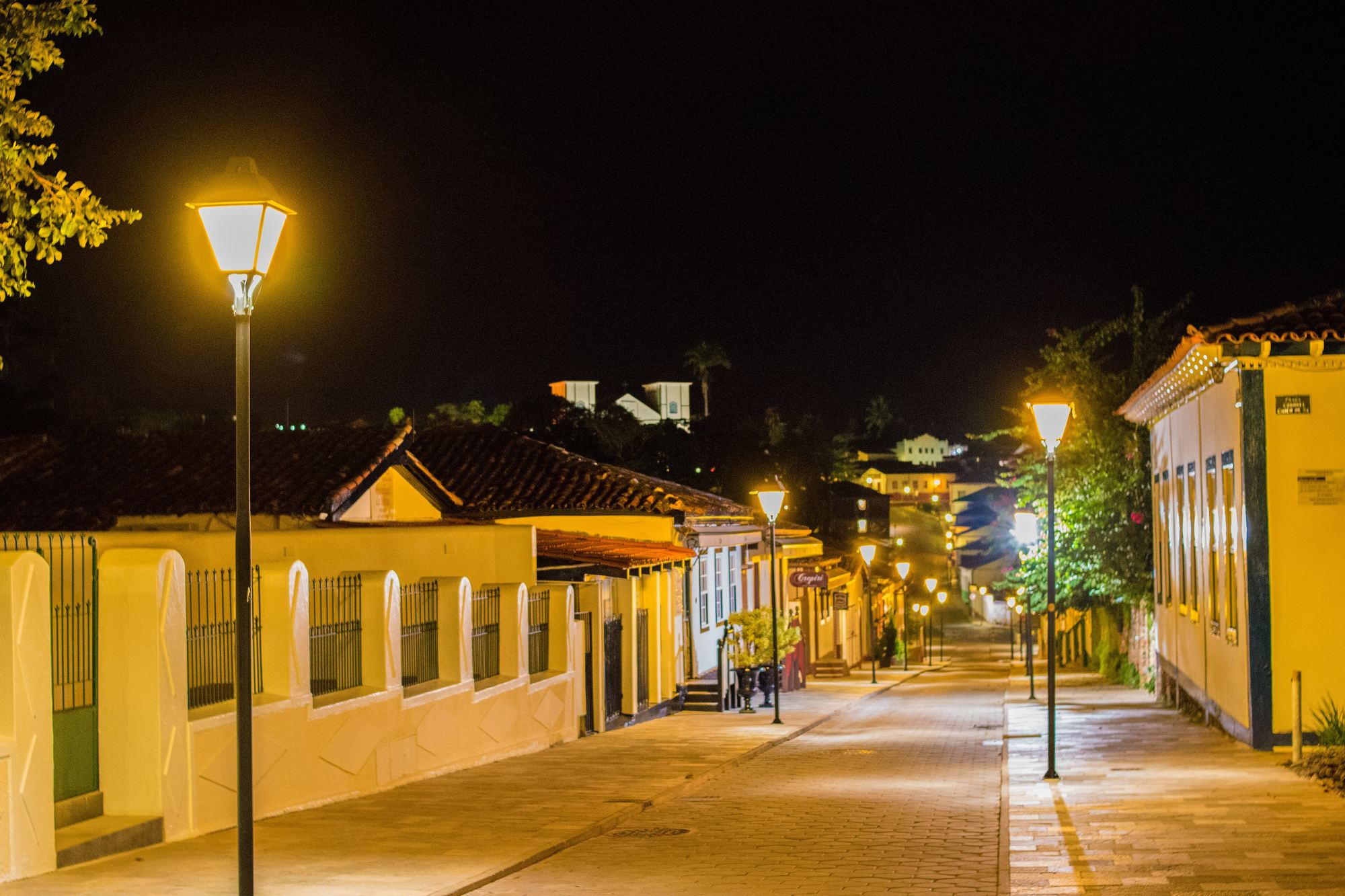 Rua do Rosário, a Rua do Lazer de Pirenópolis, à noite, com calçamento de pedra, postes acesos, casarões coloniais amarelos e brancos dos dois lados e as torres da Igreja Matriz iluminadas ao fundo.