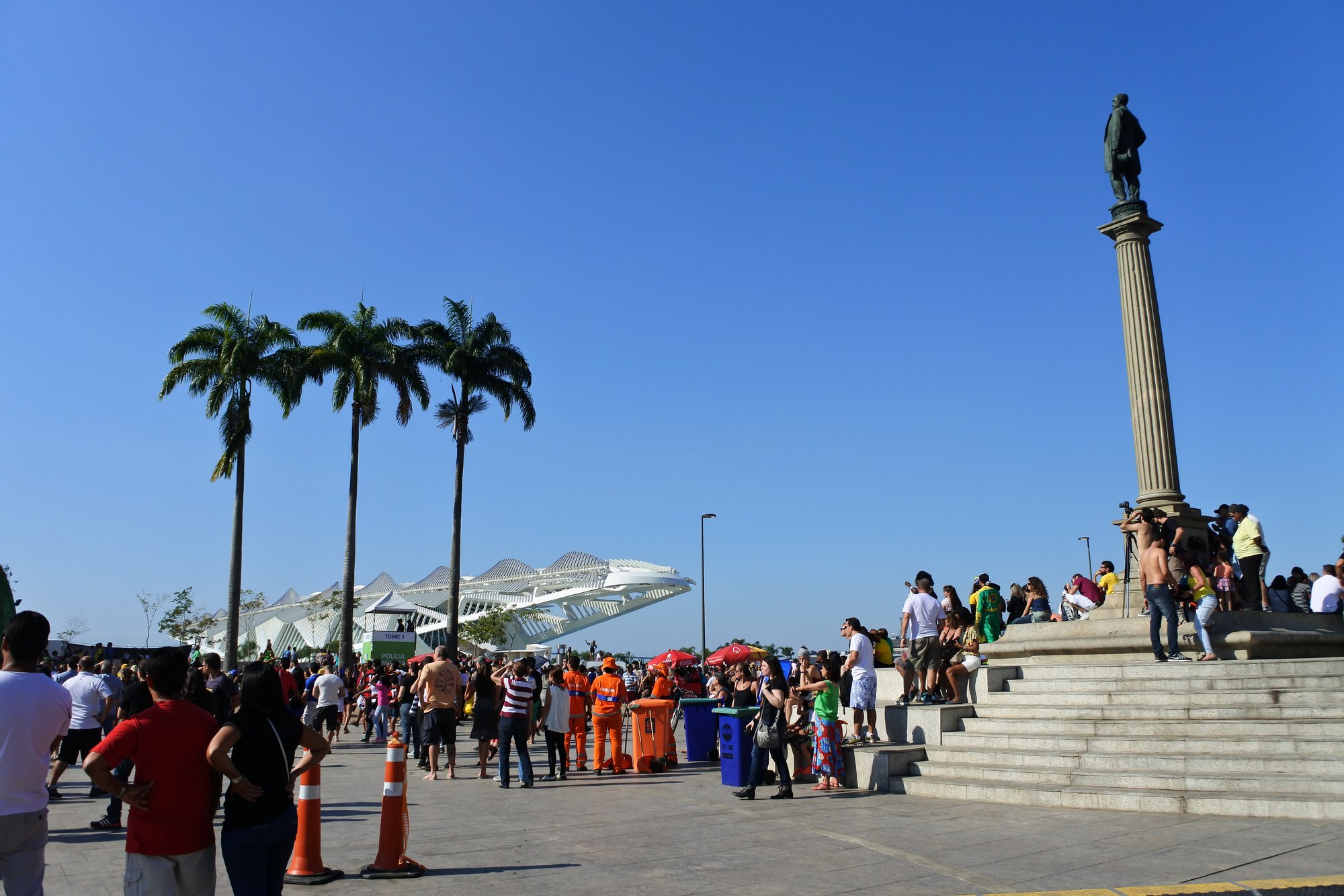 Público caminhando e sentado na escadaria da Praça Mauá, no Centro do Rio de Janeiro, num dia de céu azul, com palmeiras e o Museu do Amanhã ao fundo.