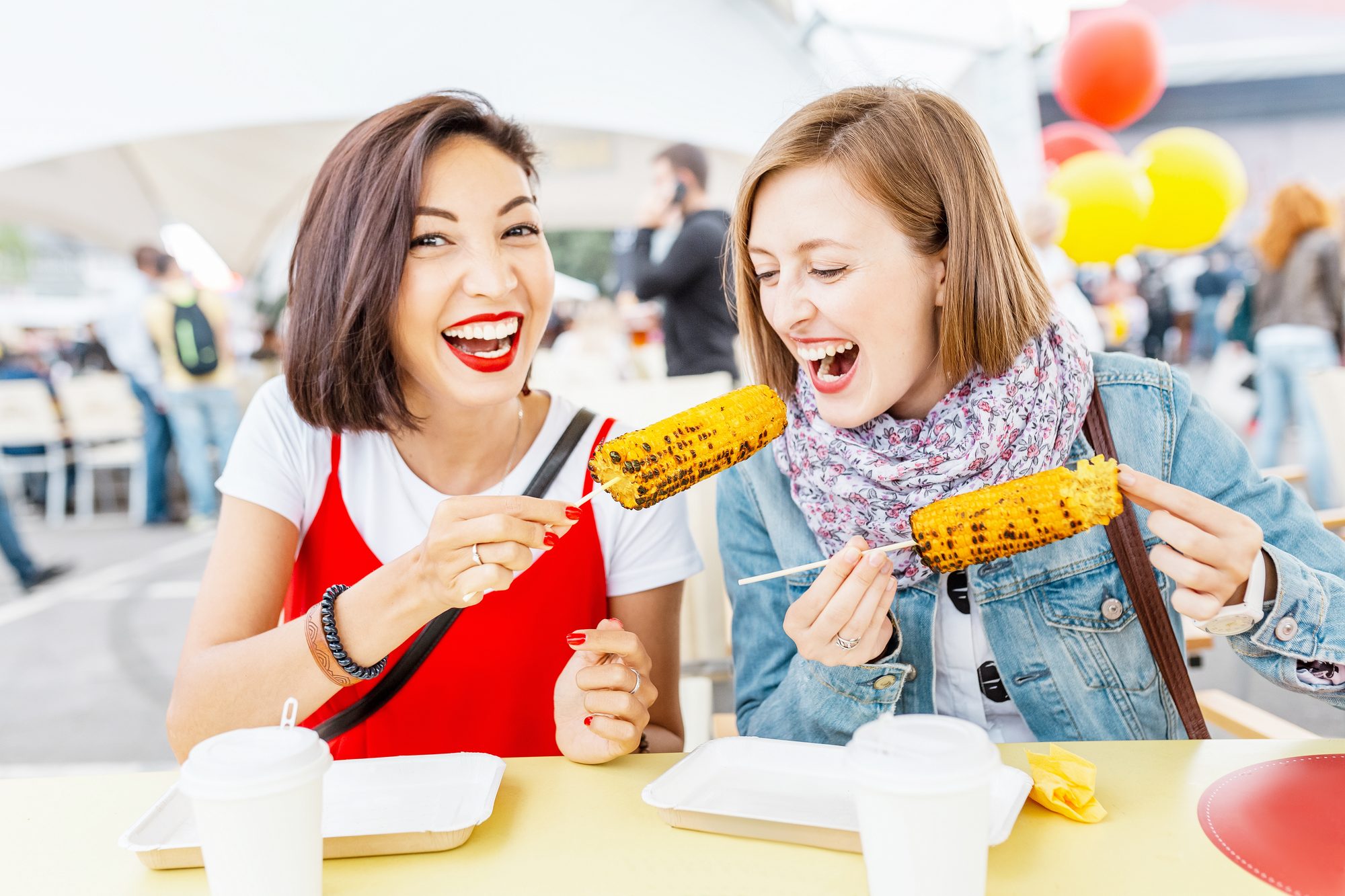 Duas amigas rindo e comendo milho assado no espeto numa mesa ao ar livre de um festival de comida, com balões coloridos e gente circulando ao fundo.