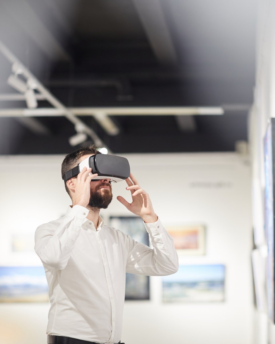 Homem de camisa branca usa óculos de realidade virtual e estende as mãos no ar numa sala de exposição, com quadros nas paredes.