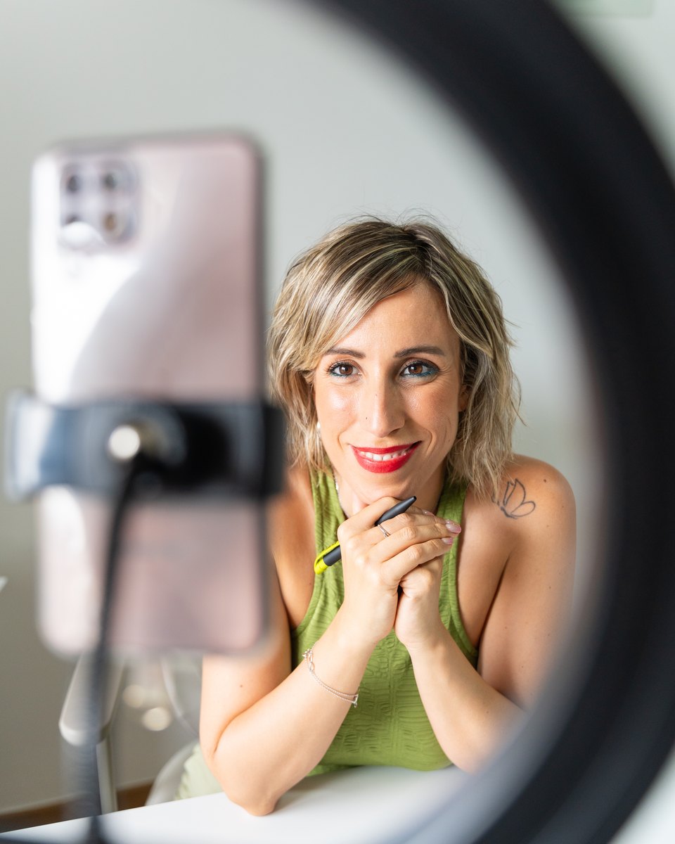 Creator sorrindo diante do celular preso num tripé, vista através de um ring light, enquanto grava um vídeo sentada à mesa.