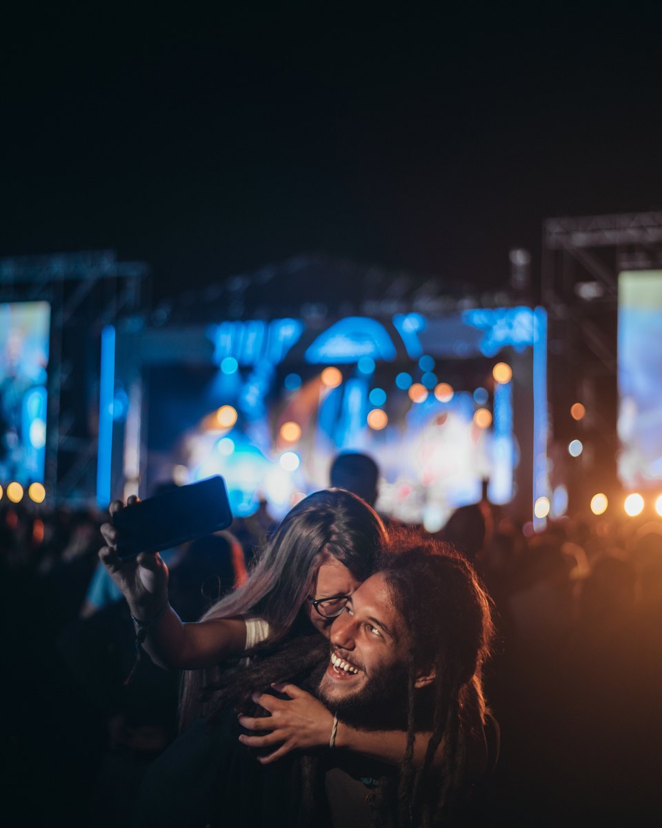 Casal abraçado fazendo selfie com o celular à noite num festival de música, com o palco iluminado de azul ao fundo.