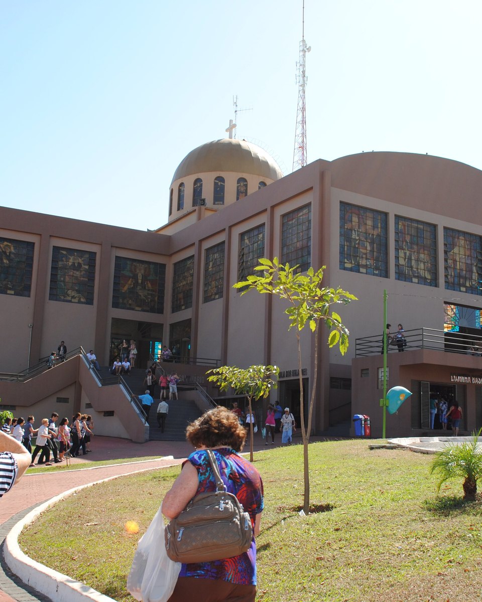 Visitantes caminhando pelo jardim em direção ao Santuário Basílica do Divino Pai Eterno, em Trindade, prédio de vitrais coloridos e cúpula dourada, com gente subindo as rampas da entrada num dia de sol.