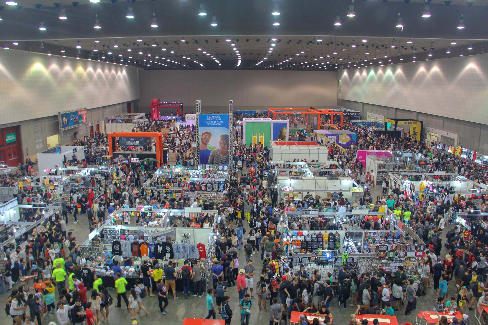 Pavilhão do Centro de Eventos do Ceará lotado durante o SANA 2018, visto do alto, com corredores de estandes de camisetas, bonecos e acessórios geek e milhares de visitantes circulando.