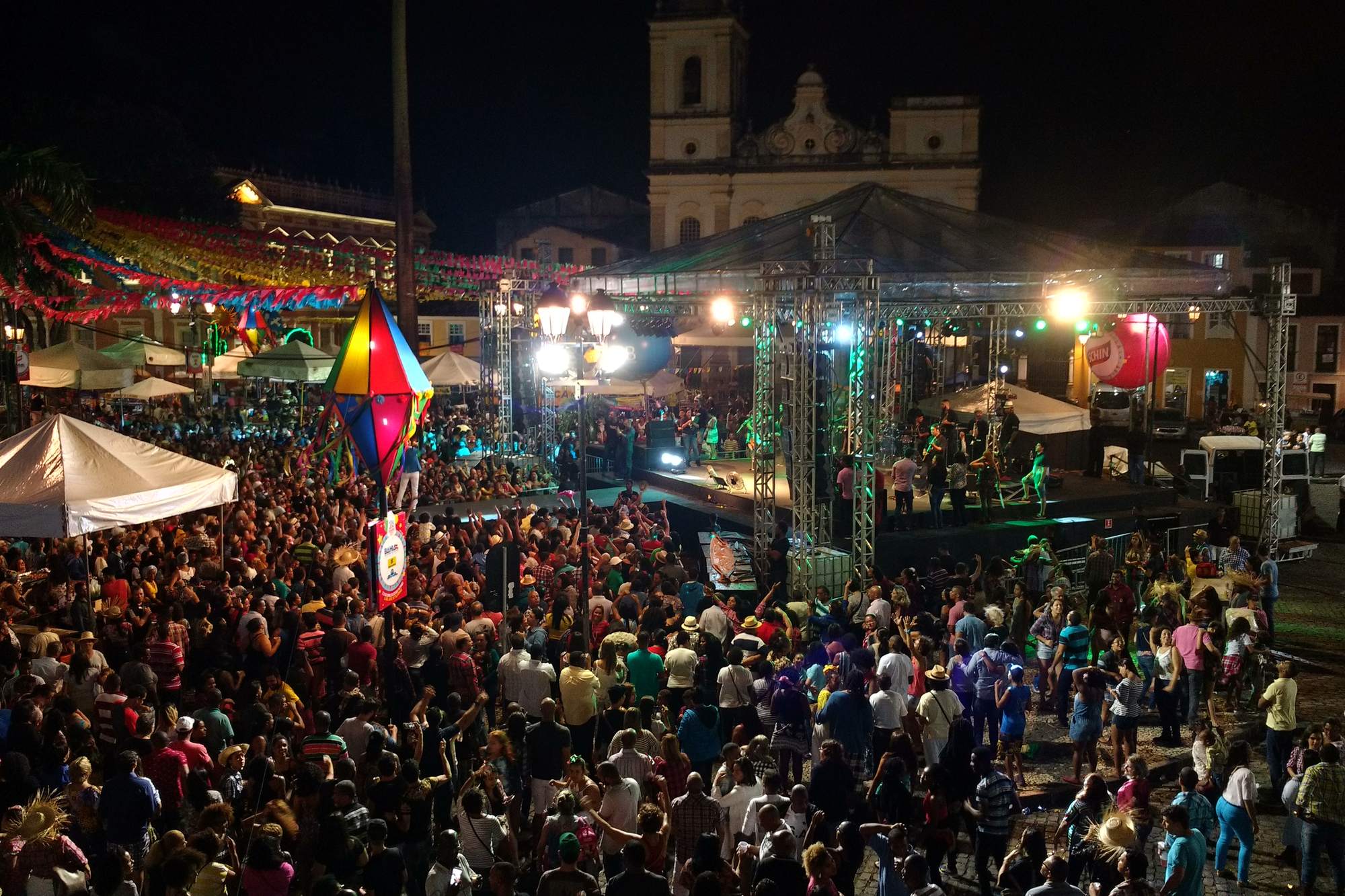 Multidão dançando numa noite de São João no Terreiro de Jesus, em Salvador, com um balão junino colorido, cordões de bandeirinhas, barracas e um palco iluminado, e as torres da Catedral Basílica ao fundo.