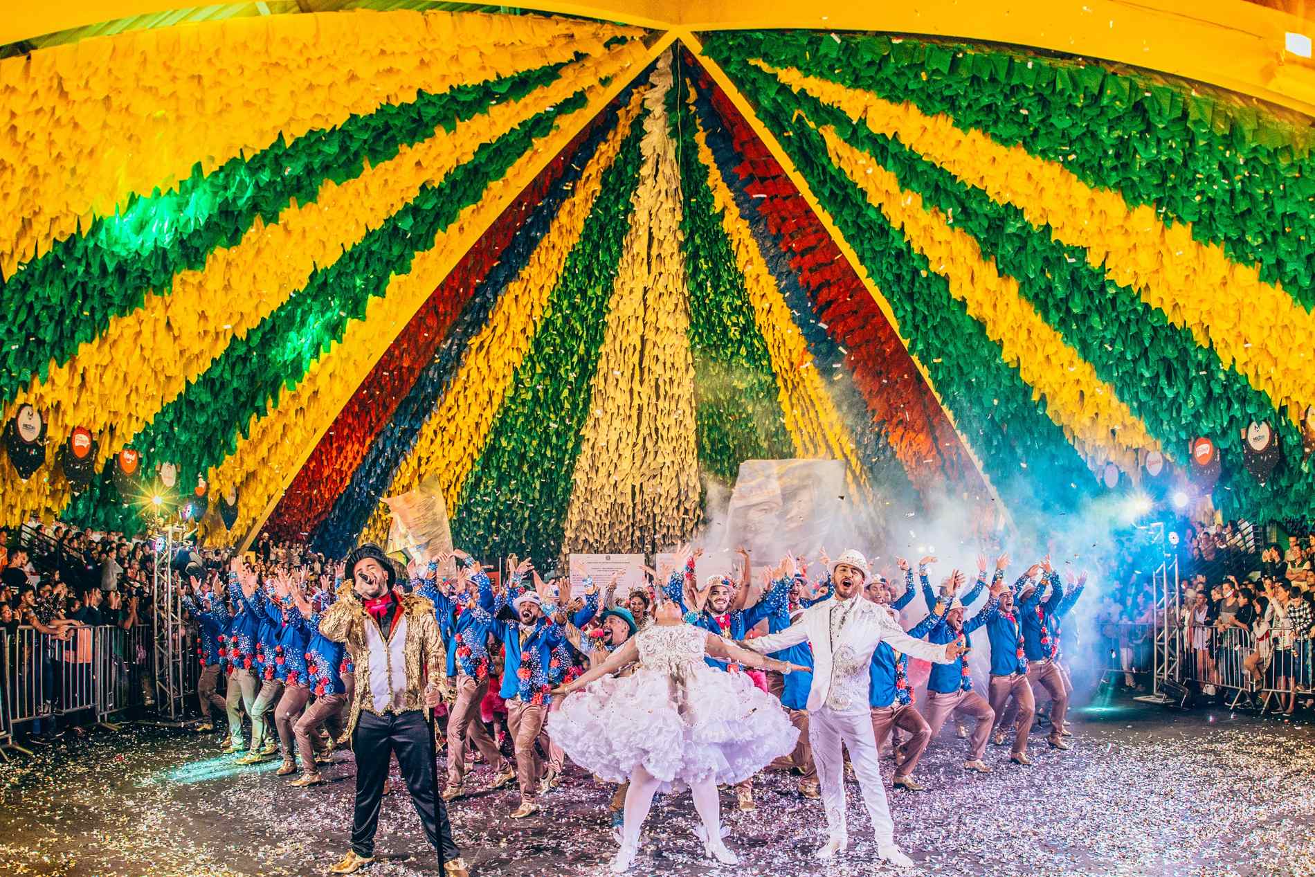 Casal de quadrilha junina dançando sob uma cobertura enorme de bandeirinhas coloridas, com o grupo em fila atrás e o público encostado nas grades, numa noite de São João em Campina Grande.