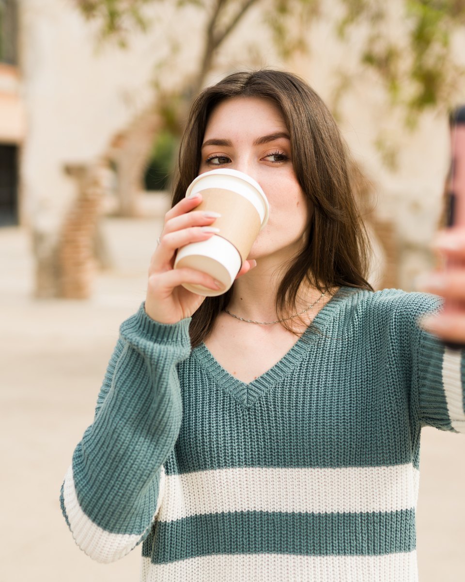 Mulher de suéter tomando café num copo para viagem enquanto se filma com o celular, numa rua arborizada.