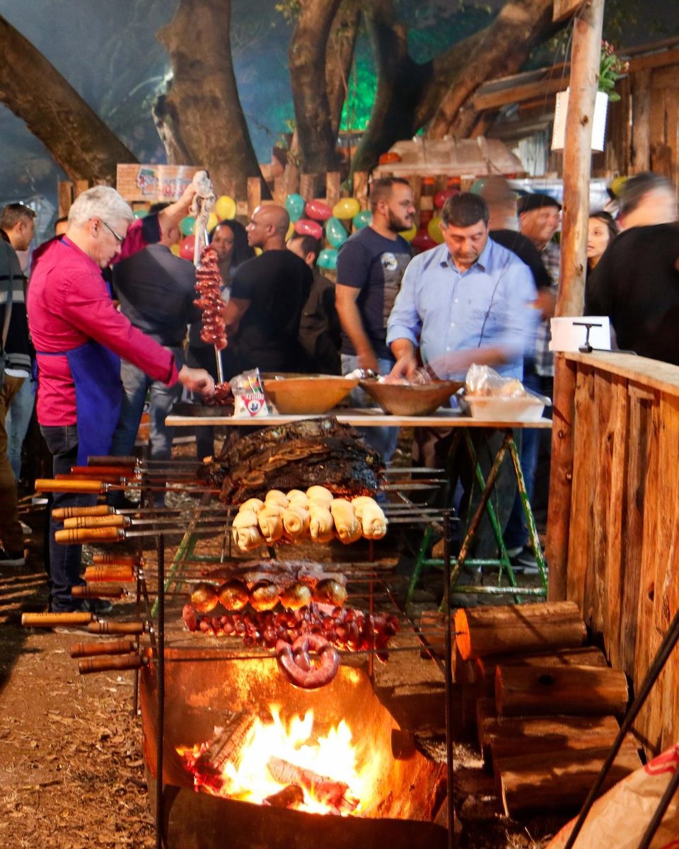Churrasco de fogo de chão num piquete do Acampamento Farroupilha, em Porto Alegre, à noite: espetos de costela, linguiça e pão sobre uma fogueira acesa num tacho de ferro no chão, um homem de avental virando o espeto e gente em volta da mesa.