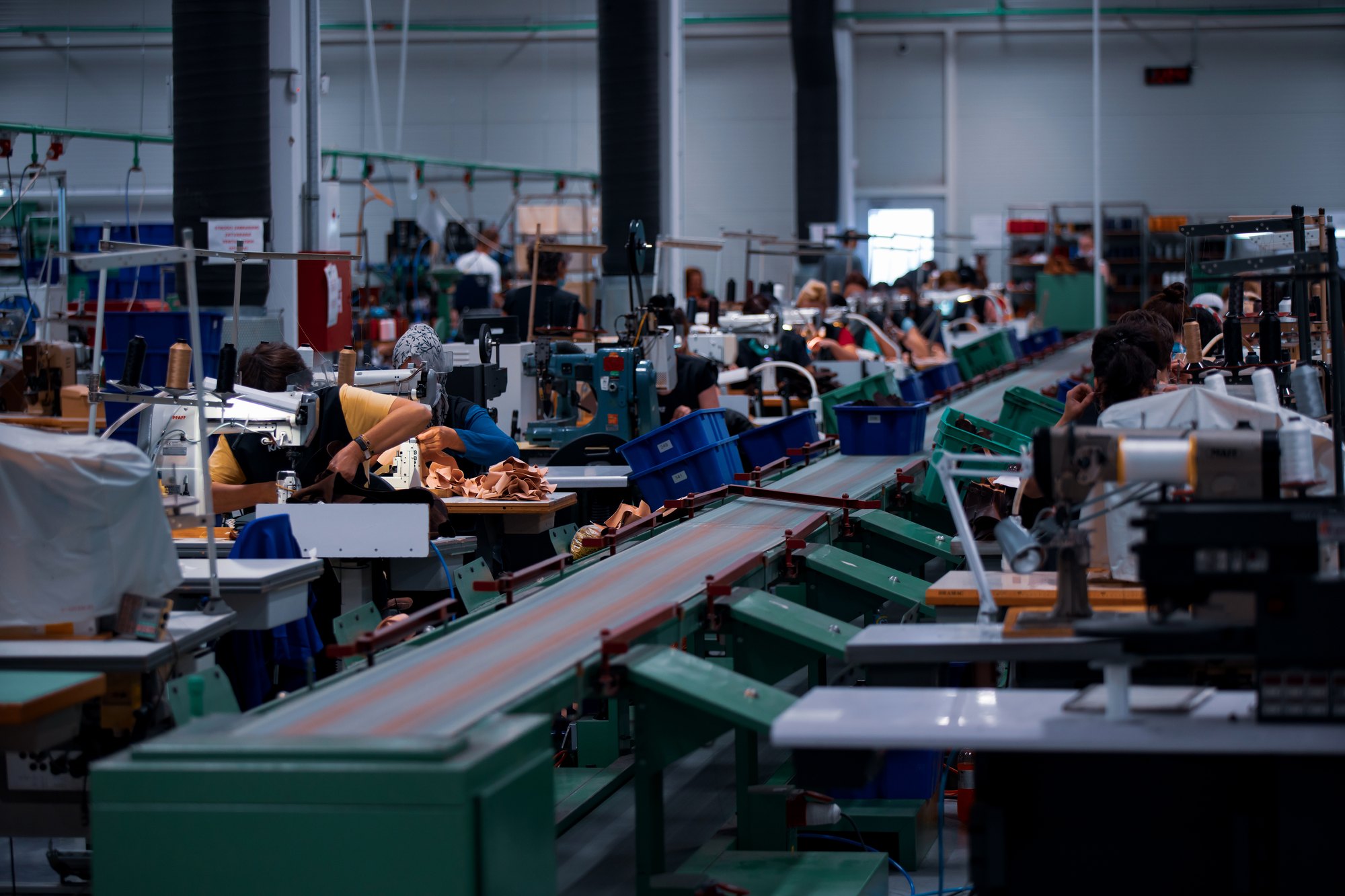 Linha de montagem de uma fábrica, com esteira verde no centro e costureiras trabalhando em máquinas dos dois lados.